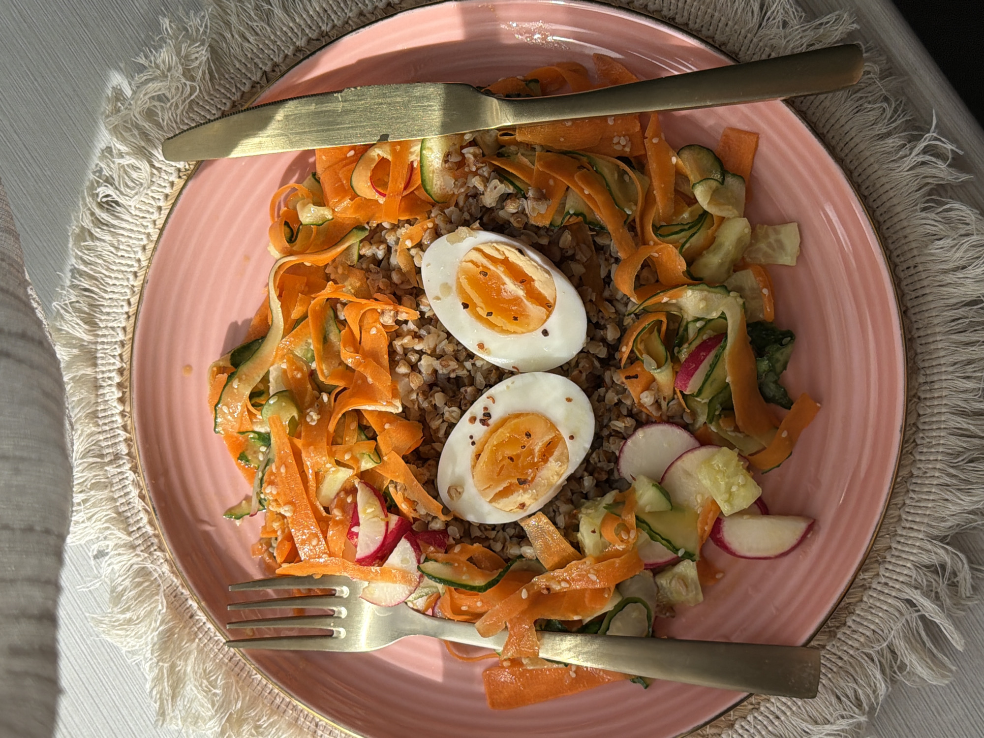 A top-down shot shows a pink plate filled with a salad consisting of buckwheat, sliced boiled eggs, and vegetable ribbons. The vegetables appear to be carrots, cucumbers, and radishes. A gold-colored knife and fork lie on top of the plate. The plate is placed on a white textured mat. The location is not identifiable as Krasnoyarsk, Russia based on the visual content of this image.