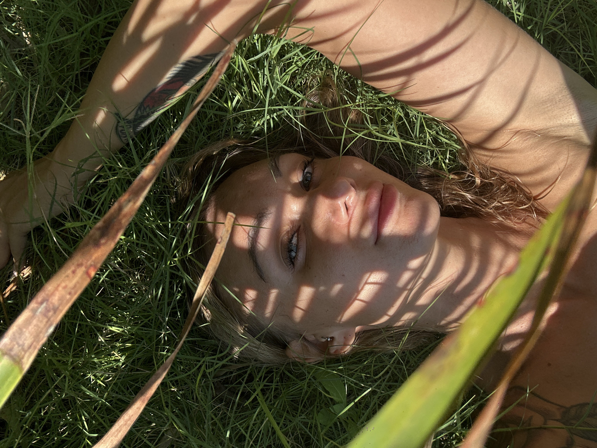 A woman lies on her back in green grass, with dappled sunlight and shadows cast across her face and body from nearby foliage. Her head is tilted slightly to her right, and she looks directly at the camera with blue eyes. She has light brown hair and fair skin. A tattoo is visible on her left arm. Blades of grass are scattered around her head and body, and sections of palm fronds partially obstruct the view. The location is Mũi Né, Vietnam.