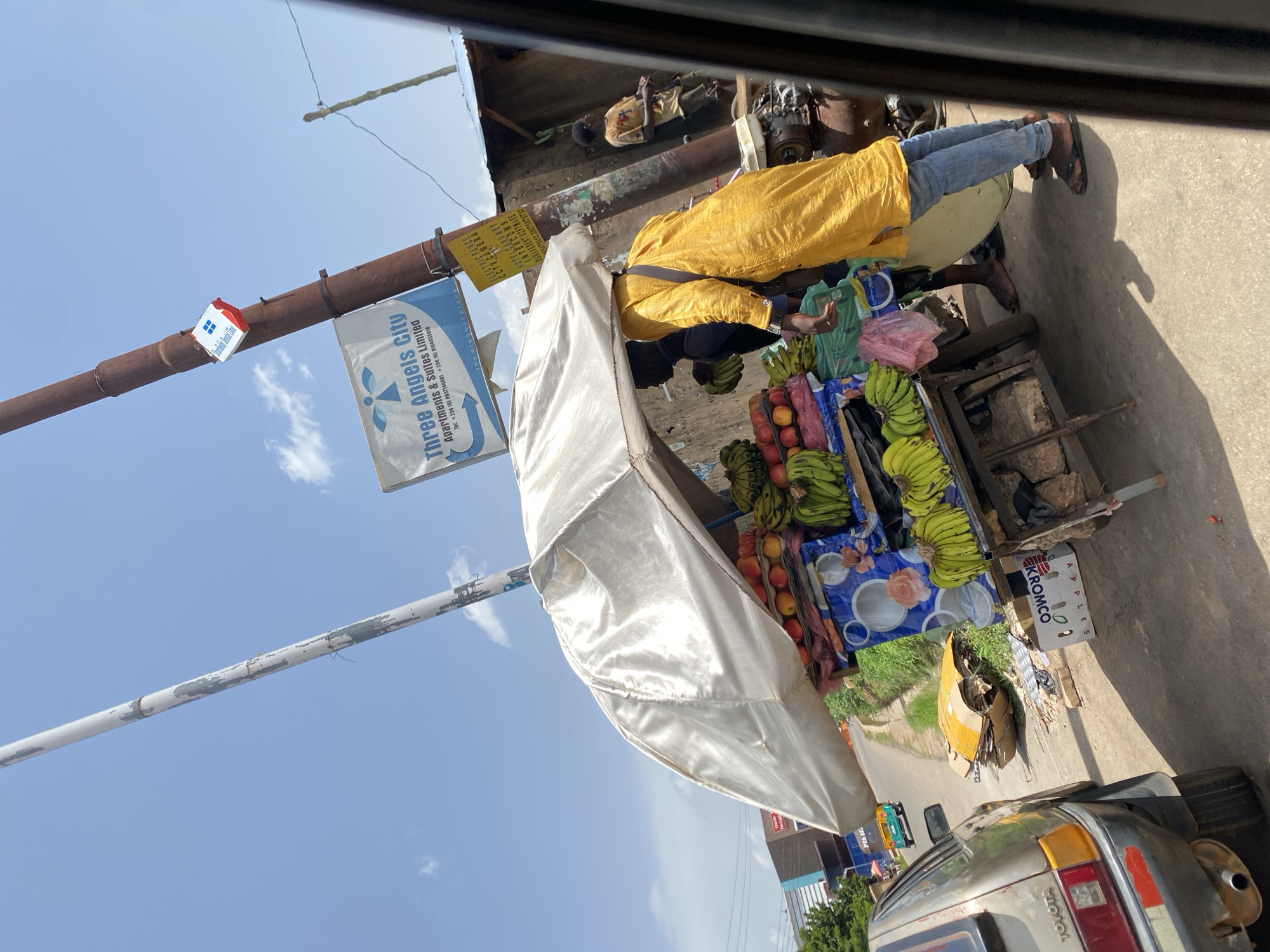The image depicts a street scene in Gura Top, Nigeria, under a bright, clear sky with scattered clouds. A fruit stall, covered by a large, shimmering white umbrella, is the central focus. The stall displays an assortment of fruits, including bunches of bananas and what appear to be apples. A man wearing a vibrant yellow robe and blue jeans stands behind the stall, seemingly attending to it or interacting with a customer. Another person is partially visible behind the stall. To the left, a light-colored Toyota car is parked, with its rear visible. In the background, various buildings and structures line the street, suggesting an urban or semi-urban setting. A pole with several signs is also prominent, one of which reads "Three Angels City Apartments & Suites Limited." The ground is paved, and there are some discarded cardboard boxes and debris near the fruit stall. The overall impression is a bustling street market scene.