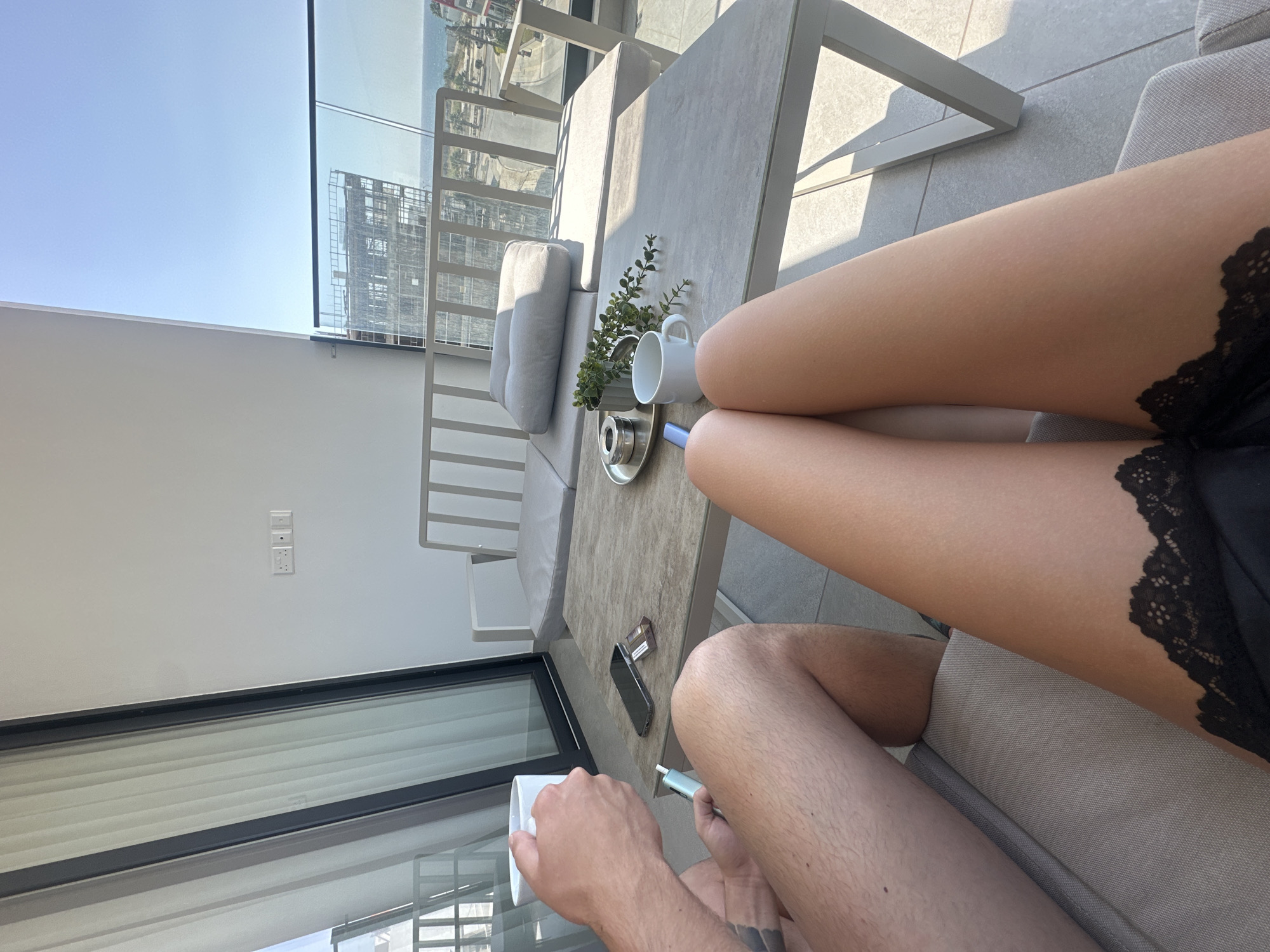 In Larnaca, Cyprus, two individuals are seated on a balcony, enjoying a sunny day. One person, whose legs are visible in the foreground, wears black lace trim underwear. The other person, on the left, holds a white mug and a light blue electronic cigarette. A modern outdoor table between them displays a smartphone, a cigarette pack, an ashtray with a cigarette, a small potted plant, and another white mug. Beyond the balcony, construction of a building is underway, and the Mediterranean Sea is visible in the distance under a clear blue sky. The scene suggests a moment of relaxation and enjoyment on a contemporary balcony overlooking the city and sea.