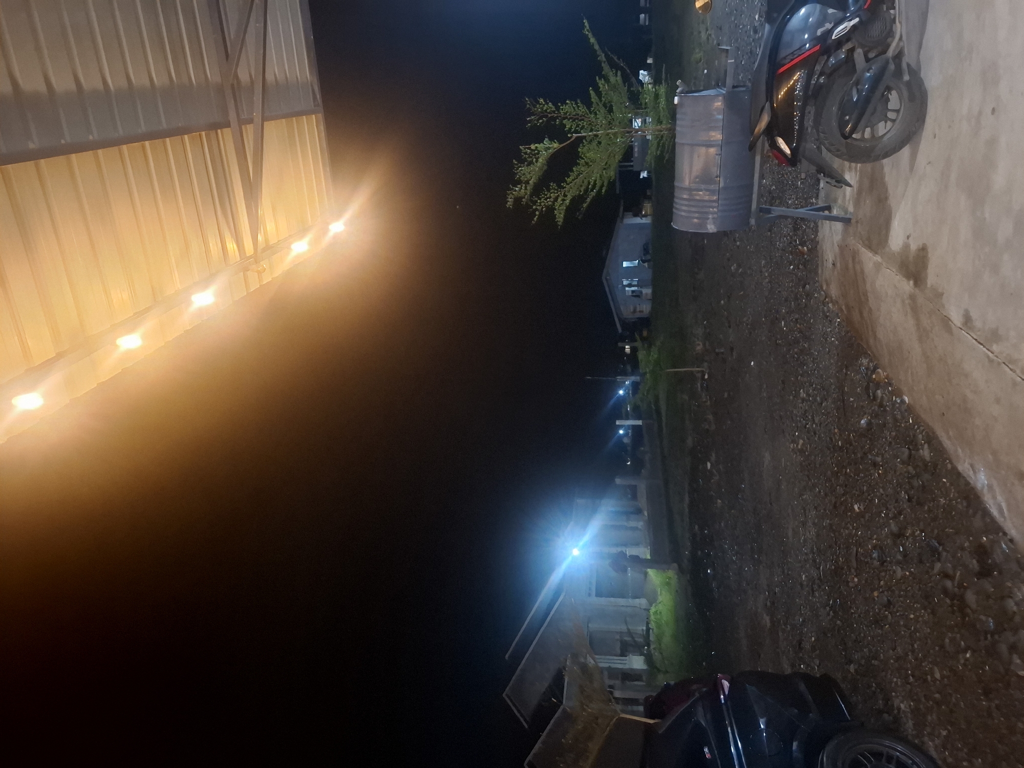 The image depicts an outdoor scene at night, with a corrugated metal roof illuminated by a string of warm-toned lights overhead. Below the roof, a dark, gravelly ground surface is visible, leading to a concrete path in the foreground. To the left, the rear portion of a black car is parked. In the mid-ground, a partially constructed building with prominent pillars and angled roof structures stands against a dark night sky, illuminated by a bright, blue-toned light source. Further back, a small, light-colored house with illuminated windows is visible, and a sparse tree with green foliage stands to its right. A large, metallic barrel, possibly a grill or storage container, is positioned on a stand to the right of the house, with a black motorcycle parked in front of it. The overall impression is of a somewhat undeveloped or construction site area in Kampongbaro, Indonesia, illuminated by artificial lighting.