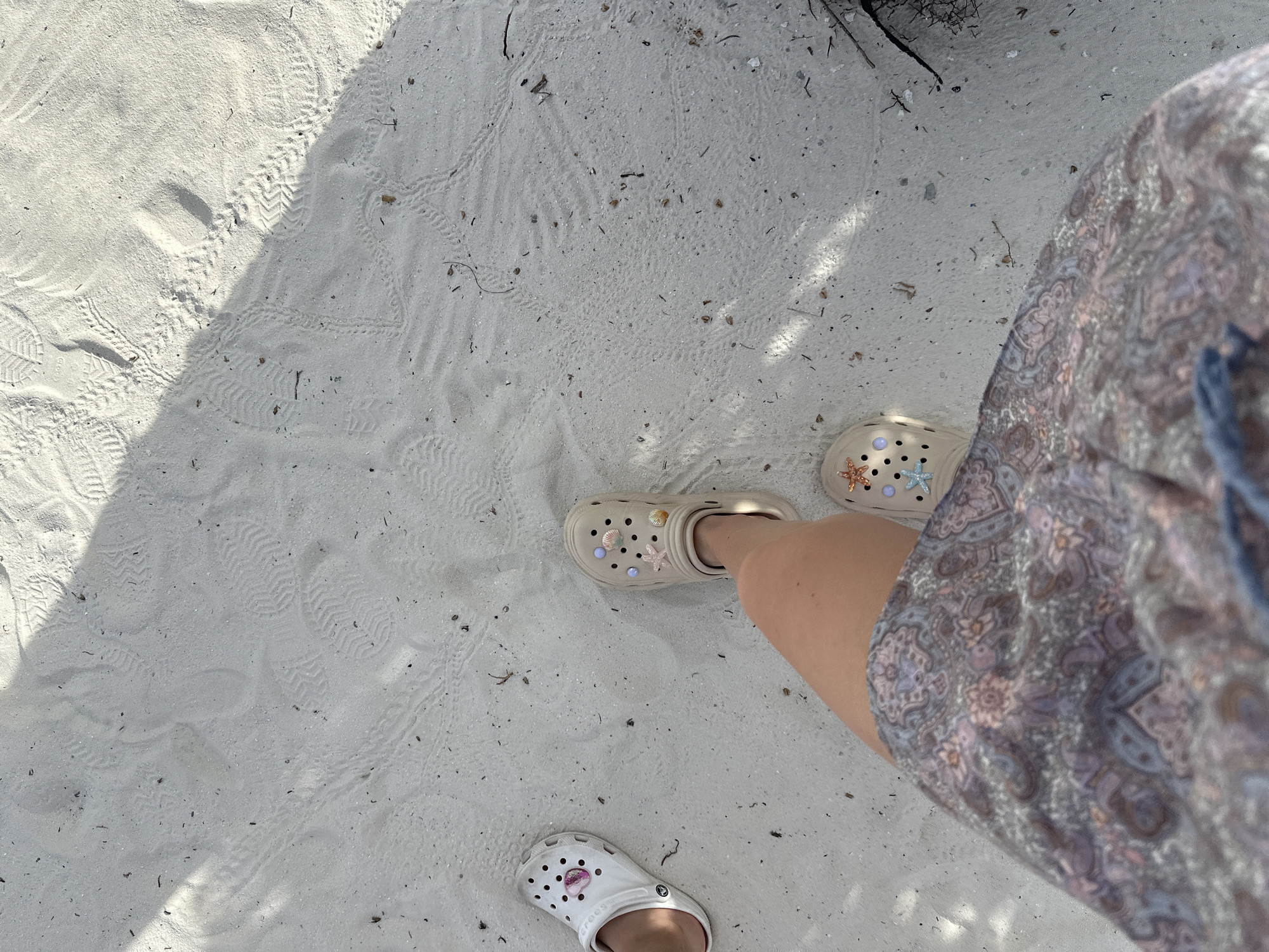 In the city of Halaveli, Maldives, a person wearing a patterned skirt and two different pairs of Crocs is standing on a sandy beach. The left foot is adorned with white Crocs, while the right foot wears beige Crocs decorated with sea-themed charms like starfish and seashells. The sandy ground displays numerous footprints and traces of what appear to be animal tracks. The scene is partially illuminated by sunlight, casting a shadow across the sand. The overall environment suggests a tropical beach setting.