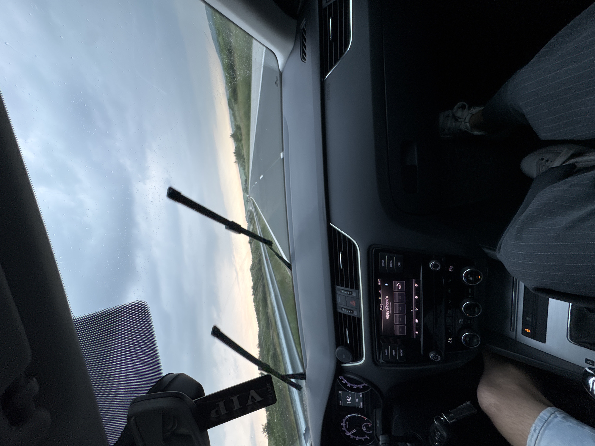 The image captures a view from inside a car, looking forward through the windshield onto a road. The scene suggests the car is in motion, driving on a highway or an open road. The sky is overcast, and water droplets are visible on the windshield, indicating recent or ongoing rain, with the car's wipers resting in an elevated position. The road ahead appears straight, flanked by guardrails and distant green landscape under a pale sky, consistent with an out-of-town drive, possibly in the Nevsha region of Bulgaria.
Inside the vehicle, a modern dashboard and central console are prominent. The infotainment screen displays the time "15:24" and "Alpay iPhone'u," suggesting a connected device, along with "SOS" and navigation/system buttons for "SETUP," "APP," and "CAR." Buttons for "RADIO," "MEDIA," and "PHONE" are also visible. Climate control settings show "19.0°C" for the left zone and "21.0°C" for the right. A partial view of the instrument cluster reveals a speed of "142" (likely km/h) and a distance reading of "1665km."
The lower legs of two occupants are visible in the cabin. On the upper right, a person is seen wearing dark, possibly pinstriped trousers and white sneakers. On the lower right, a leg clad in light-colored shorts or jeans is present, without a visible shoe. A "VIP" tag or pass is prominently hanging from the windshield on the left side, reflecting a mirrored image.