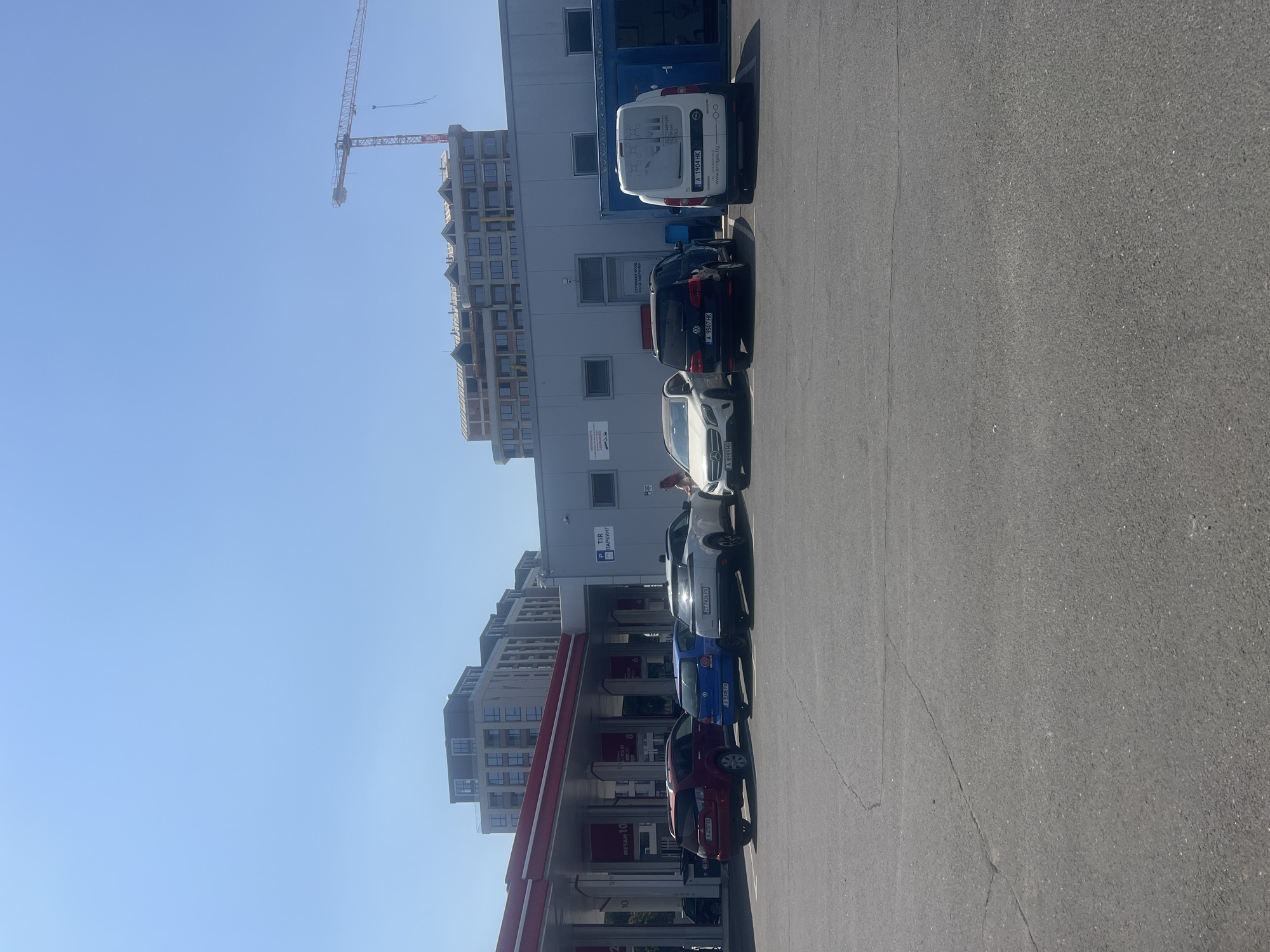 The image shows a sunny day at a gas station. A red SUV, a blue hatchback, a white hatchback, a black SUV, and a white van are parked at the gas station. In the background, there are modern buildings under construction and a large red crane. The scene appears to be in the city of Burgas, Bulgaria.