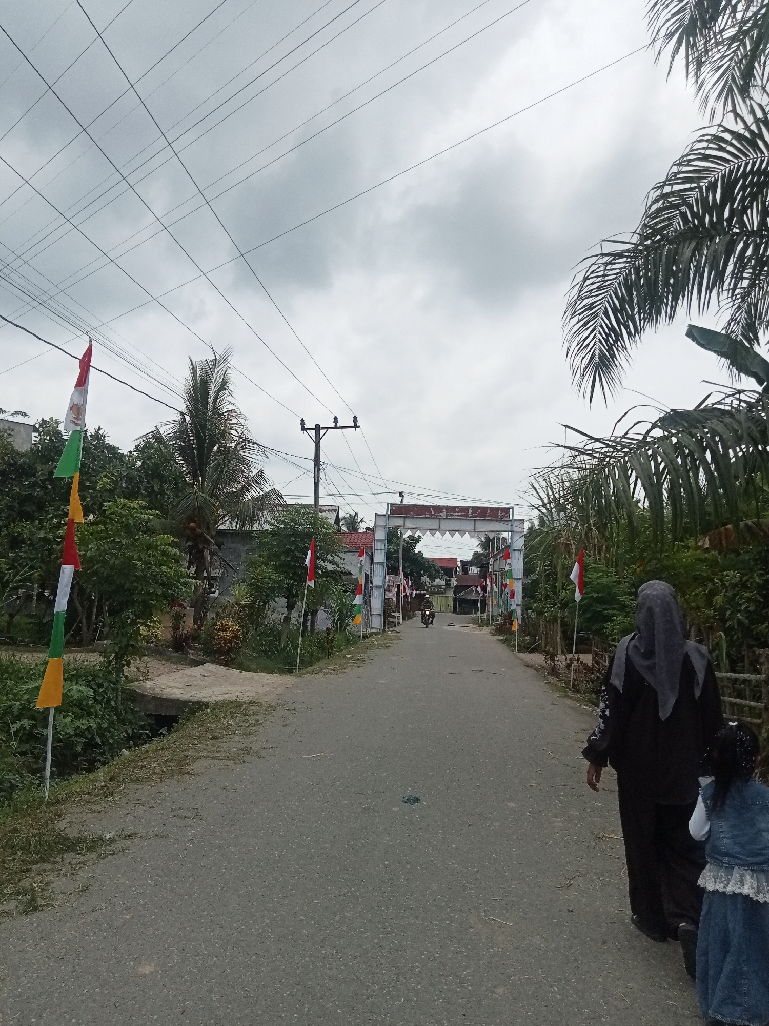 The image depicts a street scene in Singgahmata, Indonesia, on an overcast day. A narrow asphalt road leads towards a white, arched gateway decorated with triangular flags in red, white, and yellow. Numerous power lines crisscross the sky above. On the left side of the road, a series of Indonesian flags (red and white) are displayed on poles, interspersed with greenery and trees, including a palm tree. On the right side of the road, dense vegetation and more trees are visible, with some palm fronds extending into the frame. A person wearing a dark hijab and a dark outfit walks away from the camera on the right side of the road, accompanied by a young child wearing a denim vest over a white shirt and a blue skirt. In the distance, a motorcycle is seen approaching the gateway. The general environment appears to be a semi-rural or suburban area.