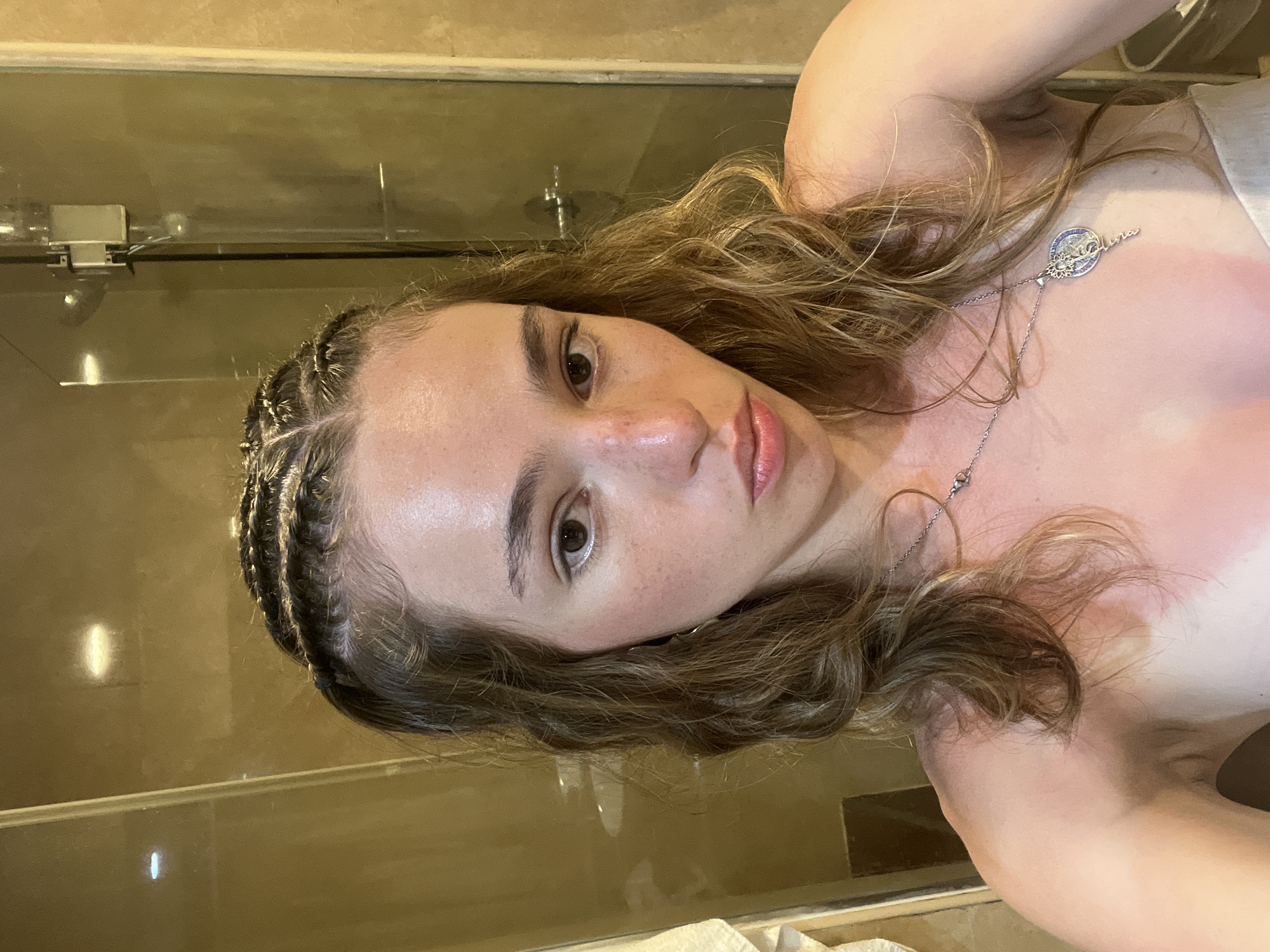 A woman is posing for a selfie indoors in a bathroom with a shower, possibly in Playa del Carmen, Mexico. She has light brown hair with dark braids along her hairline, light skin, and is wearing a silver necklace. Her skin on her chest and shoulders appears red, possibly from sunburn. The bathroom has a neutral-colored tile and glass shower door.
