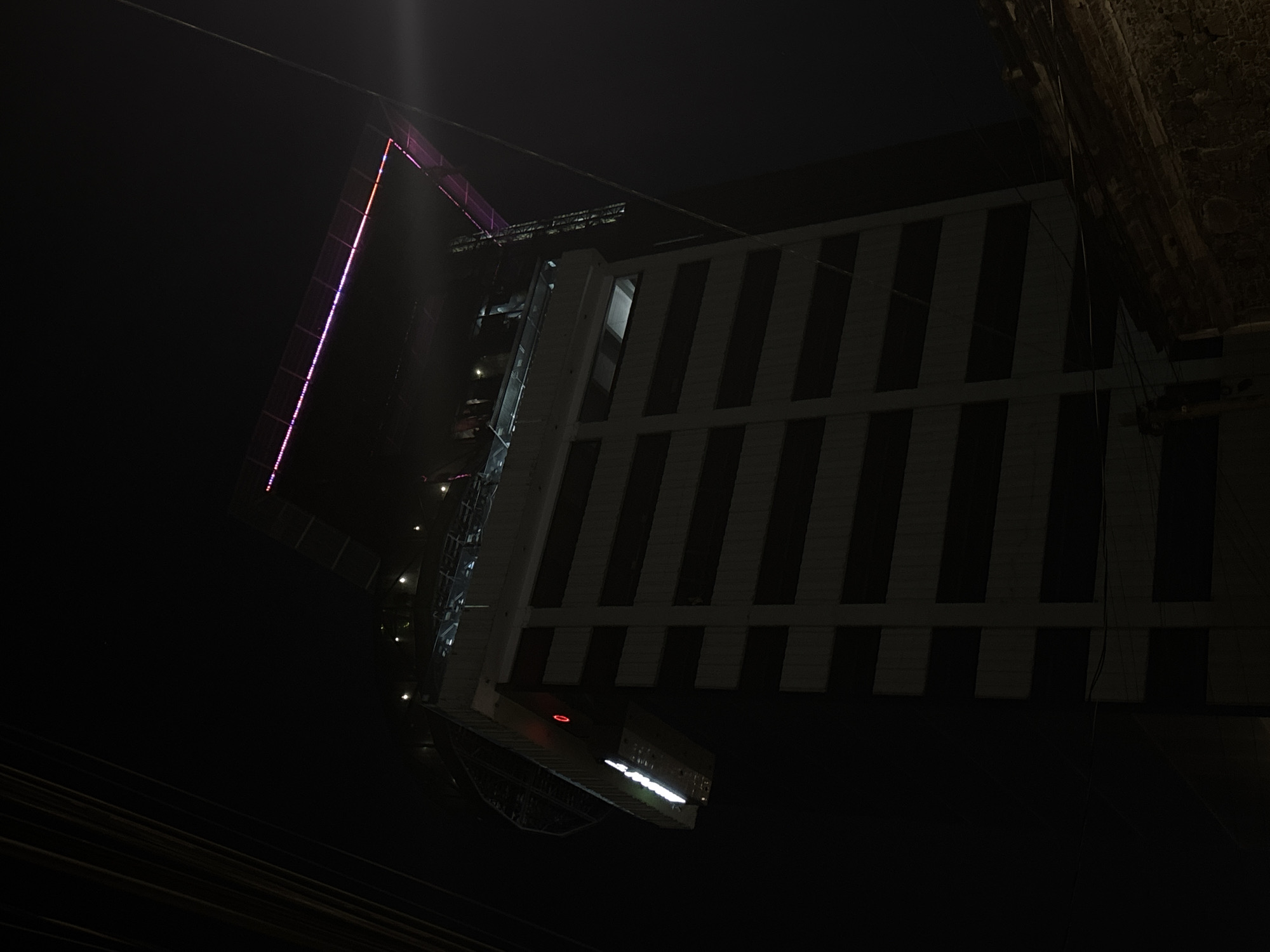 Captured at night, this low-angle shot features a tall building. The architectural structure appears modern, with alternating stripes. A thin, illuminated edge runs along one side of the building. The sky above is dark. Some cables run across the frame. The location of the image is the city of Puebla, Mexico.