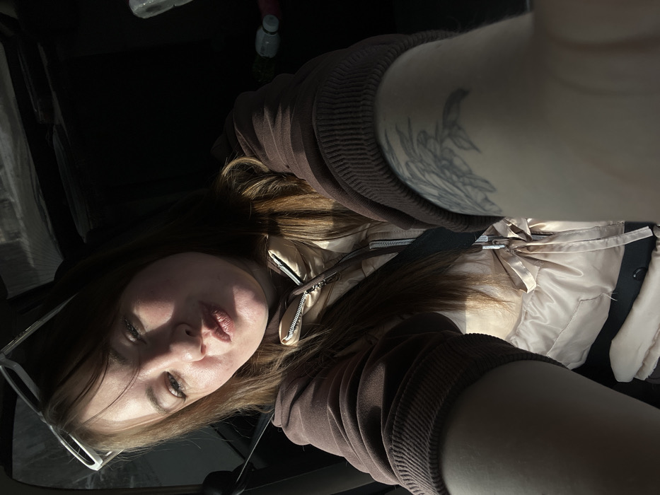 A female subject, appearing to be in her 20s to 30s with light brown hair and a floral tattoo on her left upper arm, is depicted inside a vehicle, presumably in the city of Chaikovsky, Russia. She is wearing a brown hooded sweatshirt over a light beige quilted vest and a dark seatbelt across her torso. White-framed sunglasses rest on top of her head. The subject is angled towards the camera, holding it at arm's length, with her lips pursed in a "kissing" expression. Sunlight illuminates her left cheek and forehead. The background within the car is dark, revealing indistinct shapes and two plastic bottles on a rear surface.