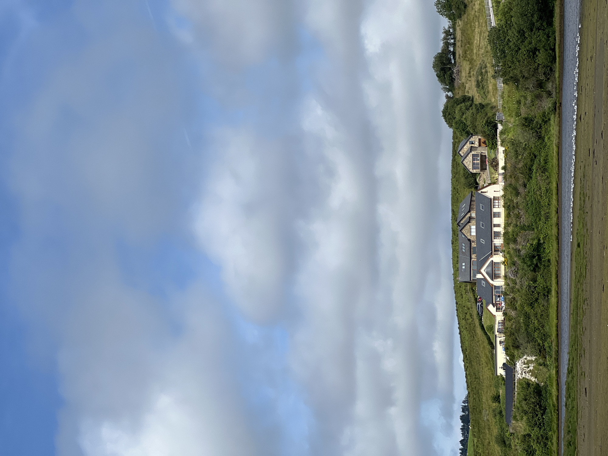 A wide, bright, daytime, full-frame, vertical shot depicts a scenic view of several modern houses situated on a grassy hill overlooking a body of water. The sky is predominantly blue with scattered, puffy, white and grey clouds. The houses are clustered together, with varying architectural styles, featuring light-colored walls, dark roofs, and numerous windows. Some of the houses have stone accents. The hill is covered in lush green grass, with scattered trees and shrubs. A white fence runs along the top of a section of the hill, and a road with a few cars is visible near the houses. In the foreground, there is a muddy, shallow body of water with a line of small, white birds on the shore. The overall impression is one of a tranquil, rural landscape in Tullycullion, Ireland, bathed in natural light.