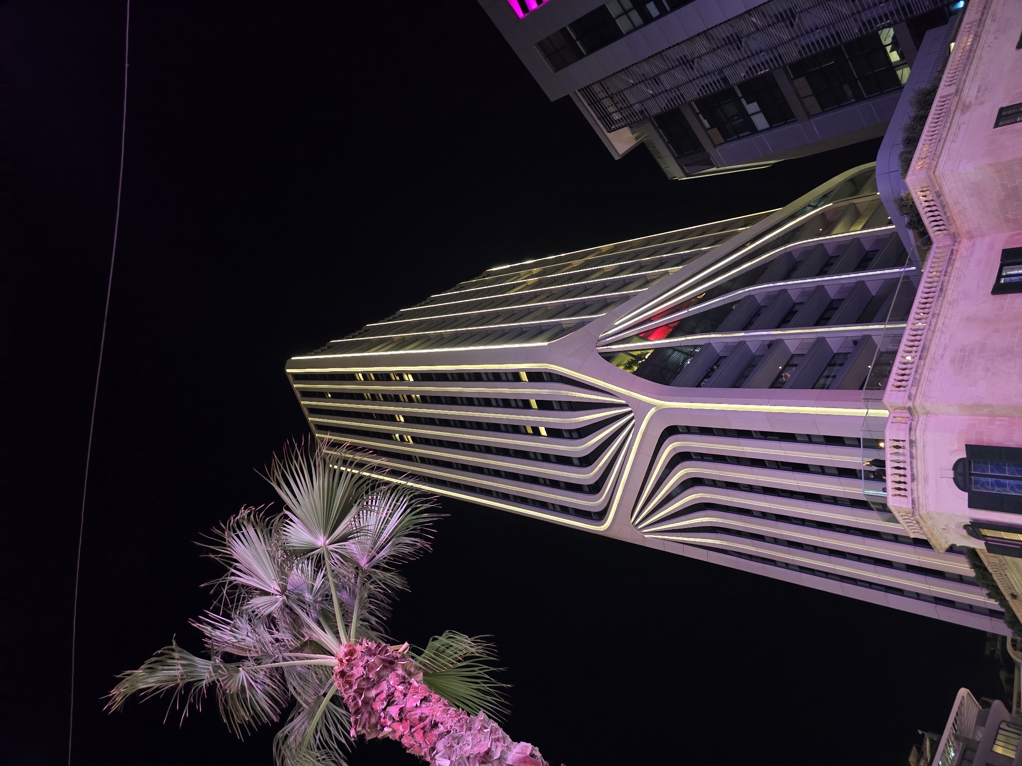 The photograph depicts a nighttime scene in Saint Julian's, Malta, focusing on a modern, illuminated skyscraper with a unique, curved facade that is outlined with bright white lights. The building's design features vertical lines that appear to fan out from the base, creating a distinctive architectural pattern. To the left of the skyscraper, a palm tree with fronds lit in pink and purple hues is visible. In the foreground, a portion of a stone building with a decorative balustrade and arched windows, also illuminated in pink, is present. Another contemporary building, partially visible to the right, displays purple lighting. The background is a dark night sky. Two individuals can be observed standing on a balcony of the lower pink building, looking towards the illuminated skyscraper.
