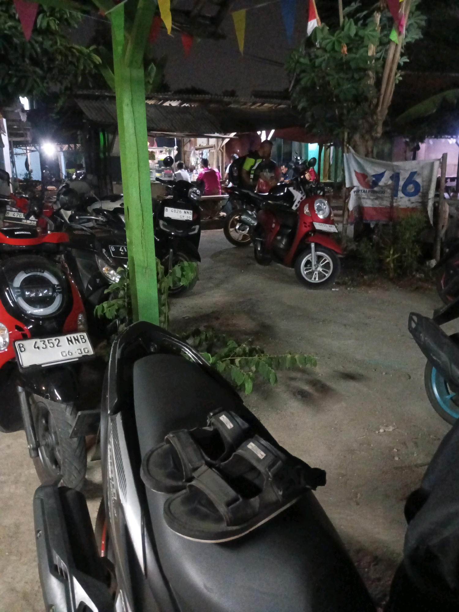 The image depicts a nighttime scene in South Tangerang, Indonesia, likely a roadside eatery or small market stall. Several parked motorcycles are visible, suggesting a casual gathering or stopping point. In the foreground, a pair of black sandals rests on the seat of a parked motorcycle. Behind the motorcycles, a food stall with customers and vendors is dimly lit. One customer is wearing a bright pink shirt. A banner with the number "16" and text is also visible. The environment is outdoors with some overhead shelter and foliage.