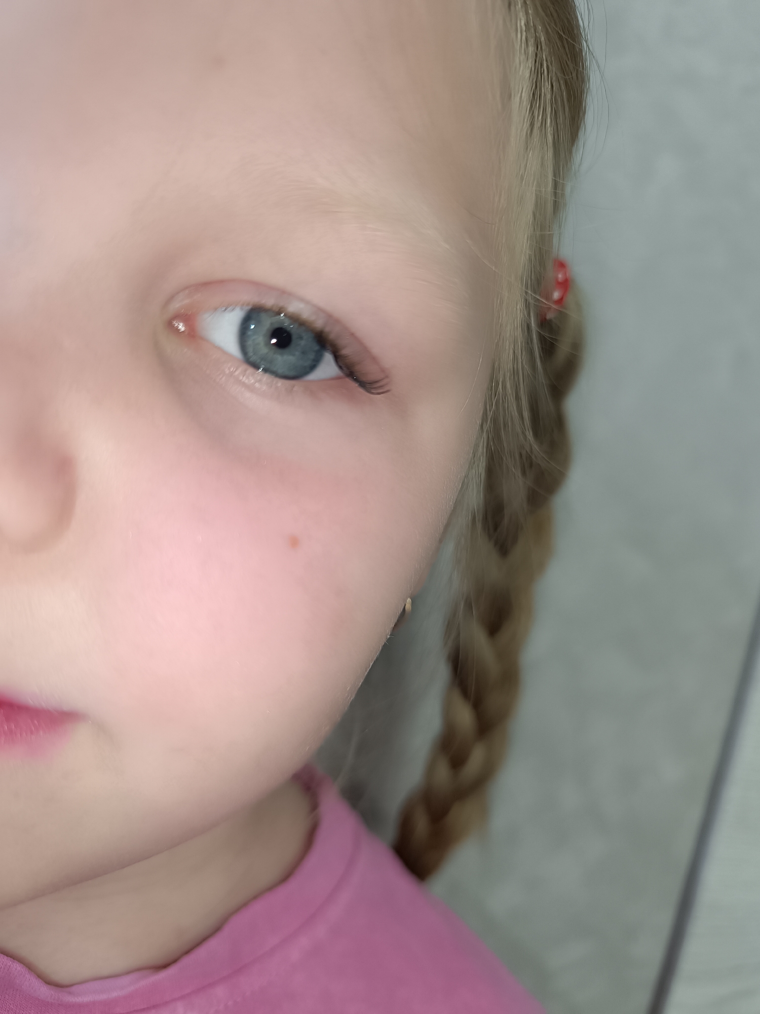 A close-up shot shows the right side of a young child's face. The child has light skin, a small brown mole on the cheek, and a blue eye with visible eyelashes. Part of the child's mouth is visible, showing pink lips. The child is wearing a pink top. Blonde hair is braided and pulled back, with a small red hair tie visible. The background is out of focus and appears to be a plain light grey or white surface. The location is Unknown, Unknown.