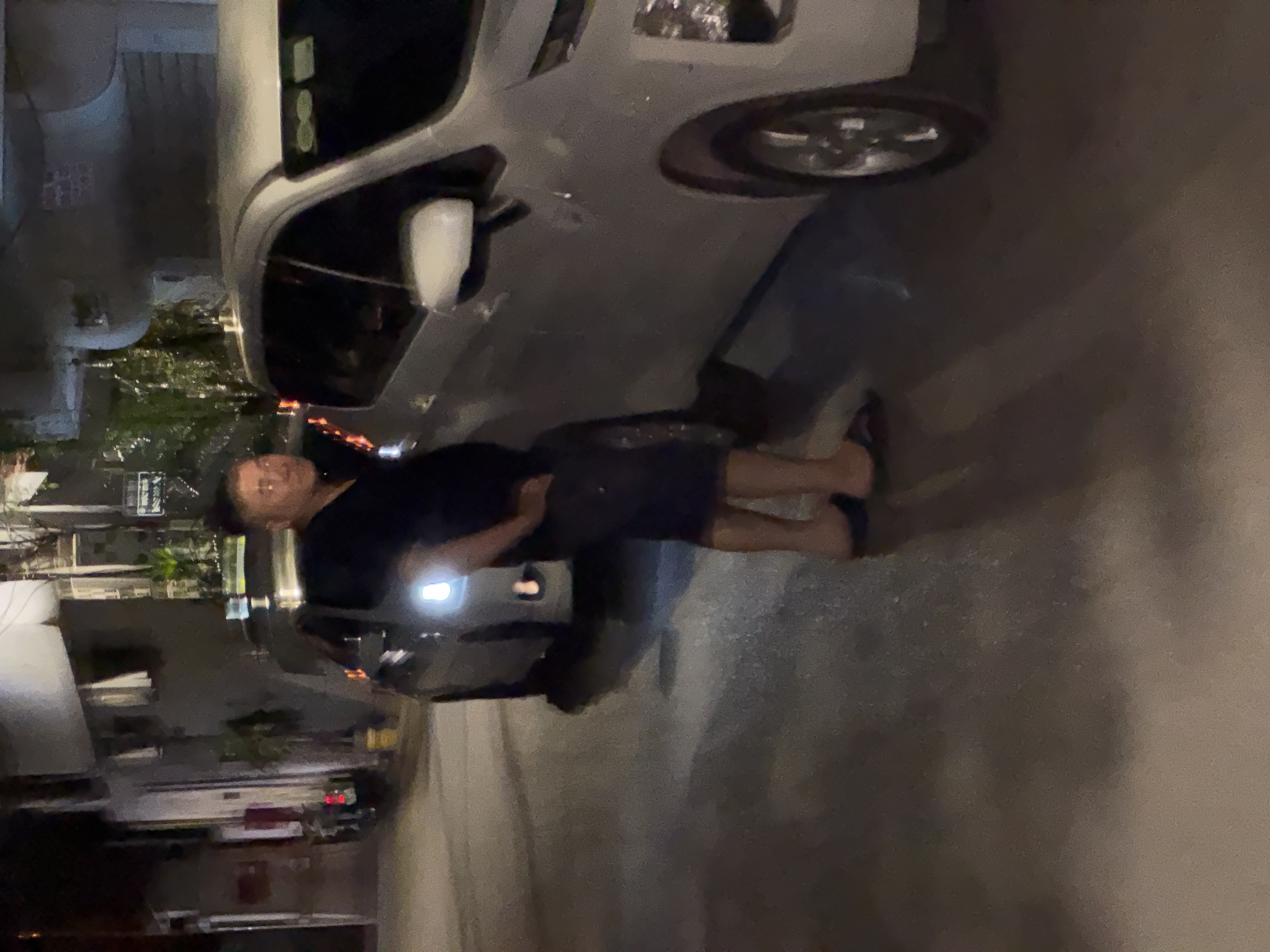 A middle-aged man in a black t-shirt and dark shorts stands on a paved street at night, leaning with his right hand on a white car. The man is facing towards the right side of the frame, looking slightly down. His left hand is resting on his hip. The car is a modern sedan with its headlights off. In the background, another car is parked, and buildings line the street, suggesting an urban setting. The street appears wet, reflecting the lights from the surrounding environment. The scene takes place in Hà Nội, Vietnam.