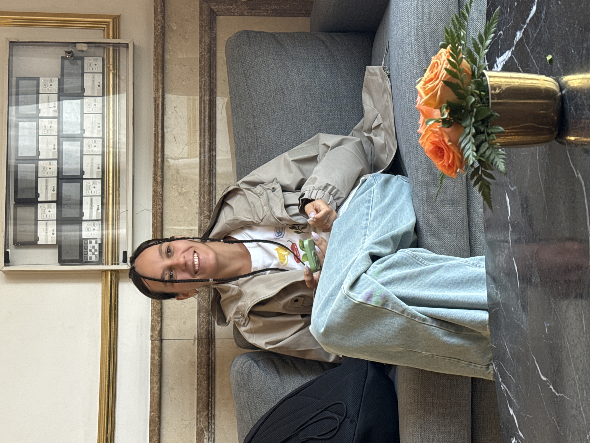 A woman sits on a gray couch next to a marble-topped table with a vase of orange roses. She has dark hair braided back from her face and is smiling at the camera. She is wearing a tan jacket and light-colored jeans. A black bag is partially visible on the couch next to her. On the wall is a framed display of small electronic devices, possibly phones, labeled "LG". The setting appears to be an indoor waiting area or lobby with patterned walls. This location is not able to be identified as Đà Nẵng, Vietnam from these details.