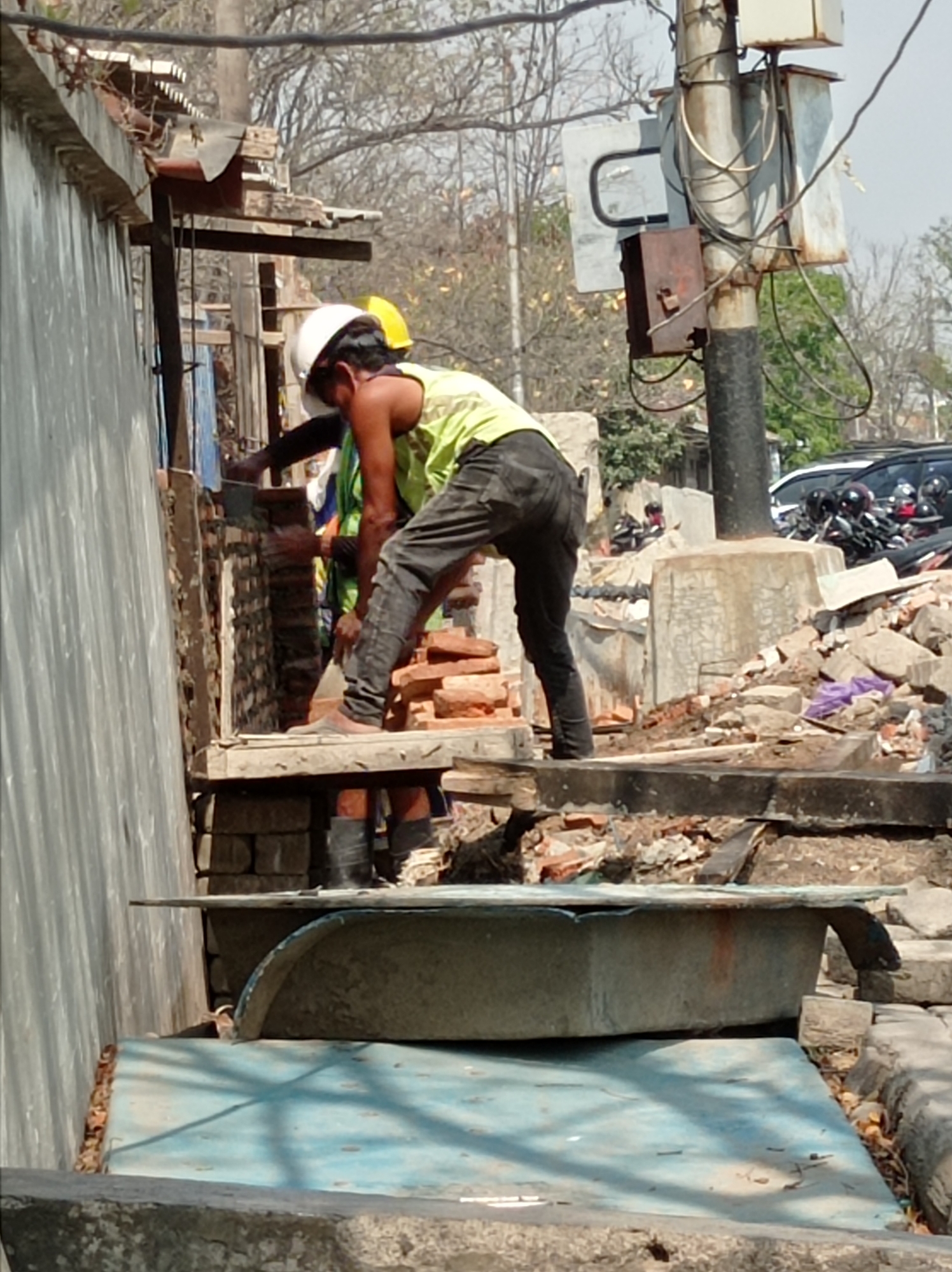 The image depicts a construction site in Tegal, Indonesia, with two construction workers in the foreground. One worker, wearing a white hard hat and a lime green vest over black pants, is actively engaged in laying bricks. He is positioned on a wooden platform, bending over a partially constructed brick wall. His right hand is holding a trowel, and his left hand appears to be placing a brick. The second worker, partially visible behind the first, is also wearing a hard hat, this one yellow. They are both wearing work boots. The background shows a damaged corrugated metal wall on the left, trees with sparse foliage, a utility pole with various electrical boxes and wiring, and a pile of rubble composed of bricks and concrete. Beyond the immediate construction area, several motorcycles and a car are parked, suggesting an urban or semi-urban setting. In the foreground, a blue tarp covers what appears to be a large, irregularly shaped object, possibly a trough or basin. The overall scene suggests ongoing repair or demolition work in a busy area.