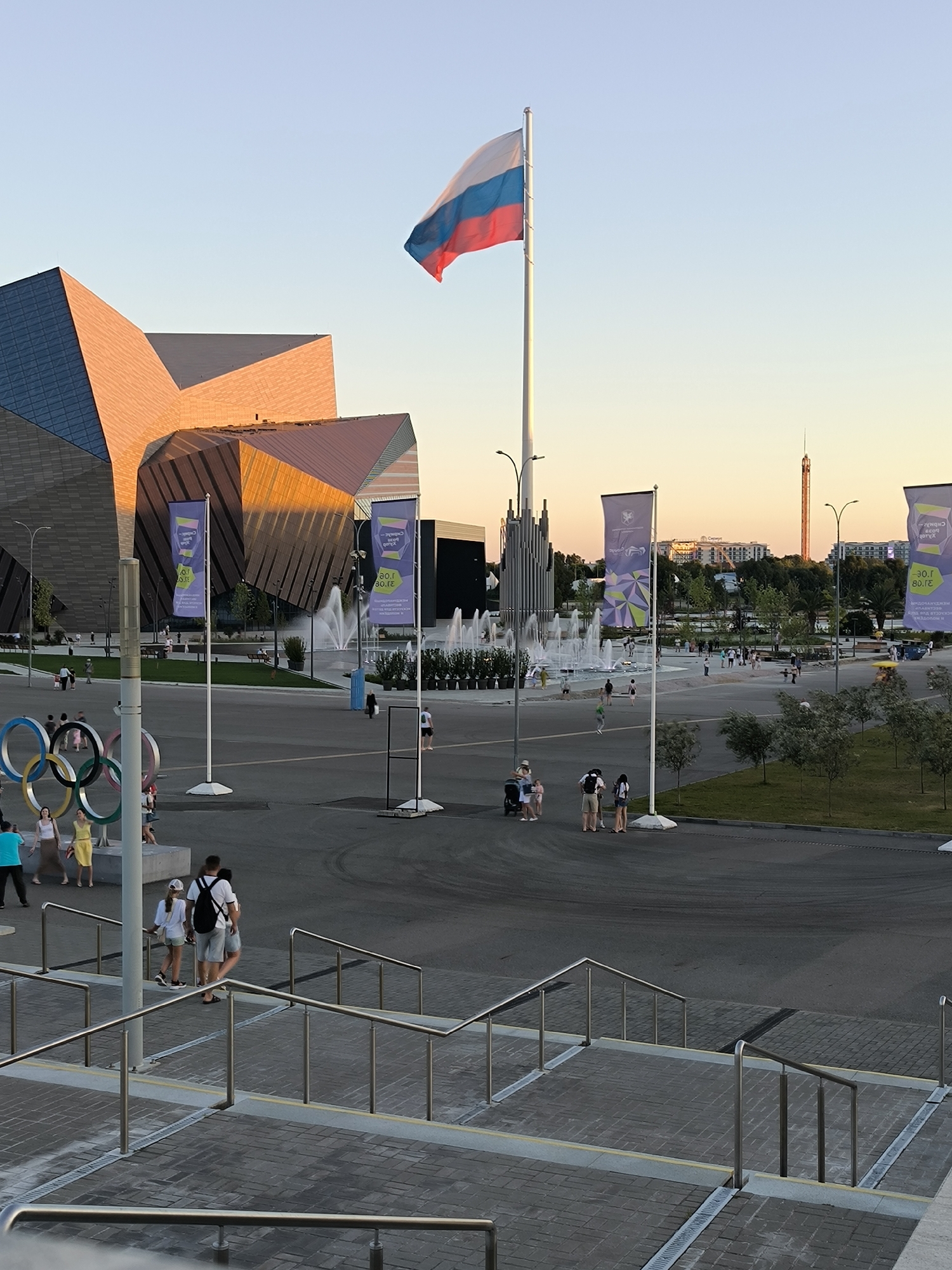 The image captures a daytime scene in the city of Sirius, Russia, likely during a warm season given the attire of the people. The focal point of the image is a large Russian flag flying on a flagpole against a clear blue sky transitioning to a pale orange hue near the horizon, indicative of either early morning or late afternoon. In the background, modern architectural structures with angular designs and metallic or glass facades are visible, reflecting the sunlight. Adjacent to these buildings, a fountain with multiple jets of water is active, creating a refreshing atmosphere. Several large banners with colorful geometric designs and text in Cyrillic are displayed on poles.
In the foreground, a set of outdoor stairs with metal railings leads down towards a paved area. On this paved area, various individuals are seen walking, standing, and interacting. Notably, a group of Olympic rings, a prominent symbol of the Olympic Games, stands to the left of the frame. The ground is a mix of asphalt and paved surfaces, with some grassy areas and trees scattered throughout the scene, suggesting a park-like setting within an urban environment. The overall impression is of a public space that is open and accessible, likely a tourist attraction or a venue for public events. The presence of people dressed casually, some pushing strollers, indicates a relaxed and leisurely atmosphere. The lighting suggests it is either sunrise or sunset, casting a warm glow on the buildings.