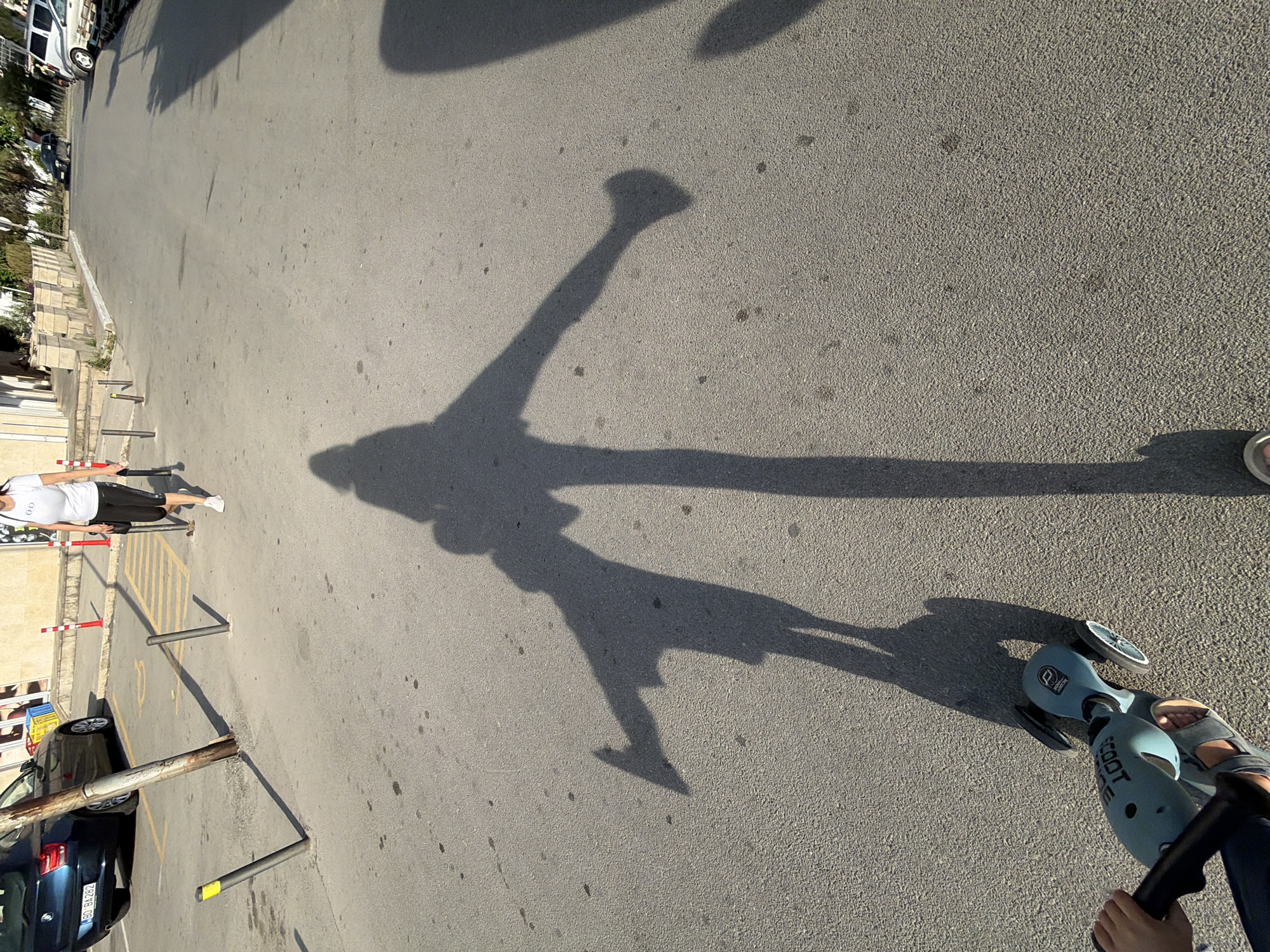 A high-angle photograph shows a paved surface, likely a parking lot or street in the city of Budva, Montenegro. A long, distorted shadow of a person is cast across the pavement. The shadow suggests the person is walking or standing, possibly with arms outstretched. To the right of the shadow, a light blue scooter is visible, with a foot in a sandal resting on it and a hand holding the handlebar. A person in a white top and dark pants is walking in the background. A row of bollards and a parked car are also present in the lower-left corner of the image.