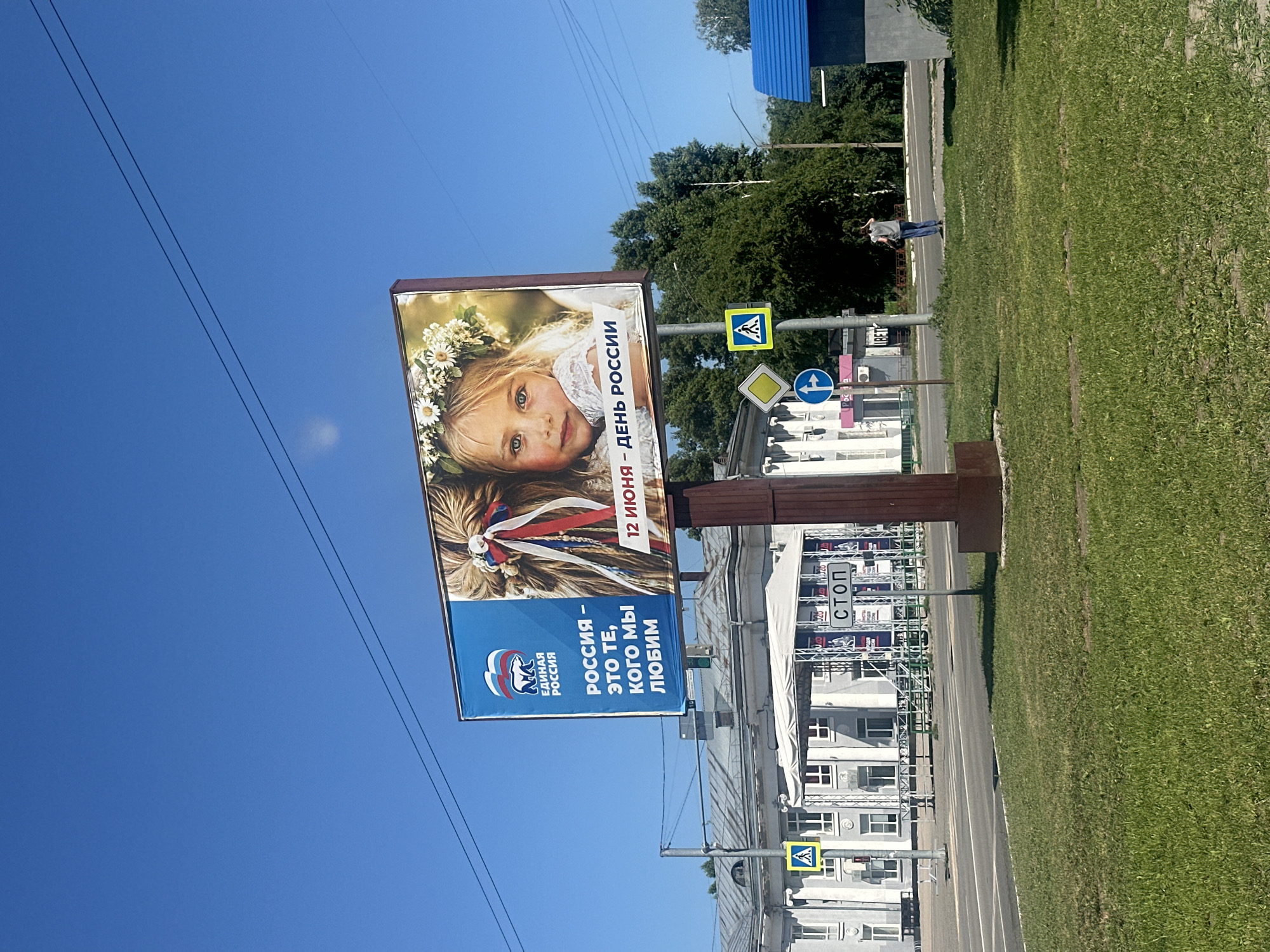 The image shows a billboard in the city of Novoaltaysk, Russia. The billboard features an image of a young girl wearing a flower crown and a quote in Russian that translates to "Russia is us, who we love." The billboard also includes the logo of the "United Russia" party. In the background, there is a building that appears to be a public institution or administrative building. A street with cars and pedestrians is visible in front of the building. The sky is clear and blue, indicating a sunny day. The overall scene depicts a patriotic advertisement in a public space.
