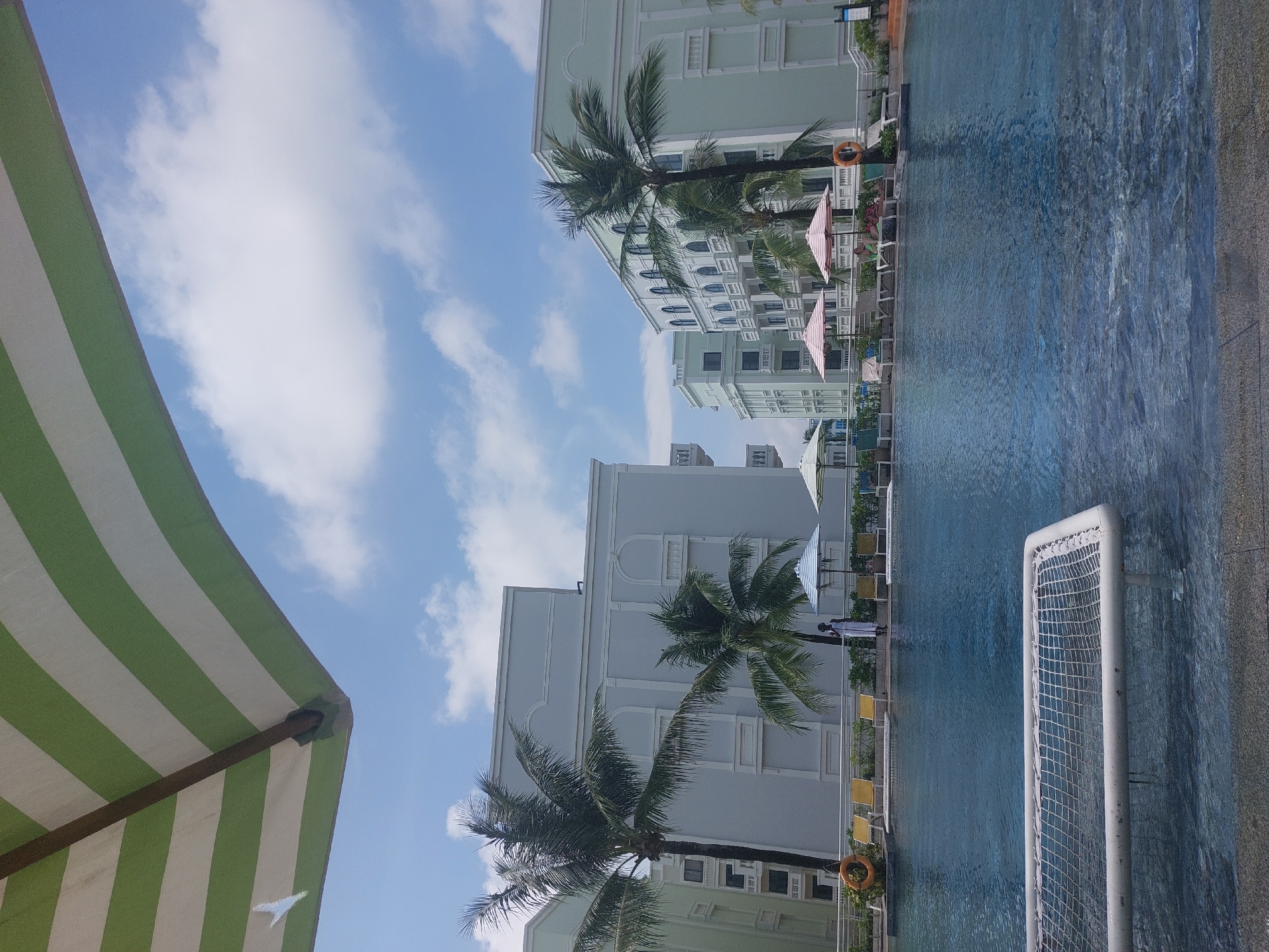 The image depicts a bright, sunny day at a resort swimming pool in Phú Quốc, Vietnam. In the foreground, the clear blue water of the pool fills the lower half of the frame. A white net-like structure, possibly part of a pool float or water feature, is visible submerged in the water near the bottom edge. Beyond the pool, a paved area features several yellow lounge chairs and a few umbrellas, one striped with pink and white.
A person stands near the edge of the pool, dressed in white. Palm trees with lush green fronds are scattered throughout the scene, with two prominent ones near the center. In the background, several multi-story resort buildings are visible. These buildings are painted in light pastel colors, primarily a pale blue-gray and a light mint green, and feature architectural details like arched windows and balconies. The sky above is a vibrant blue, dotted with fluffy white clouds. A large green and white striped umbrella is prominent in the upper left corner of the image, partially obscuring the sky. The overall impression is one of a relaxing tropical vacation setting.