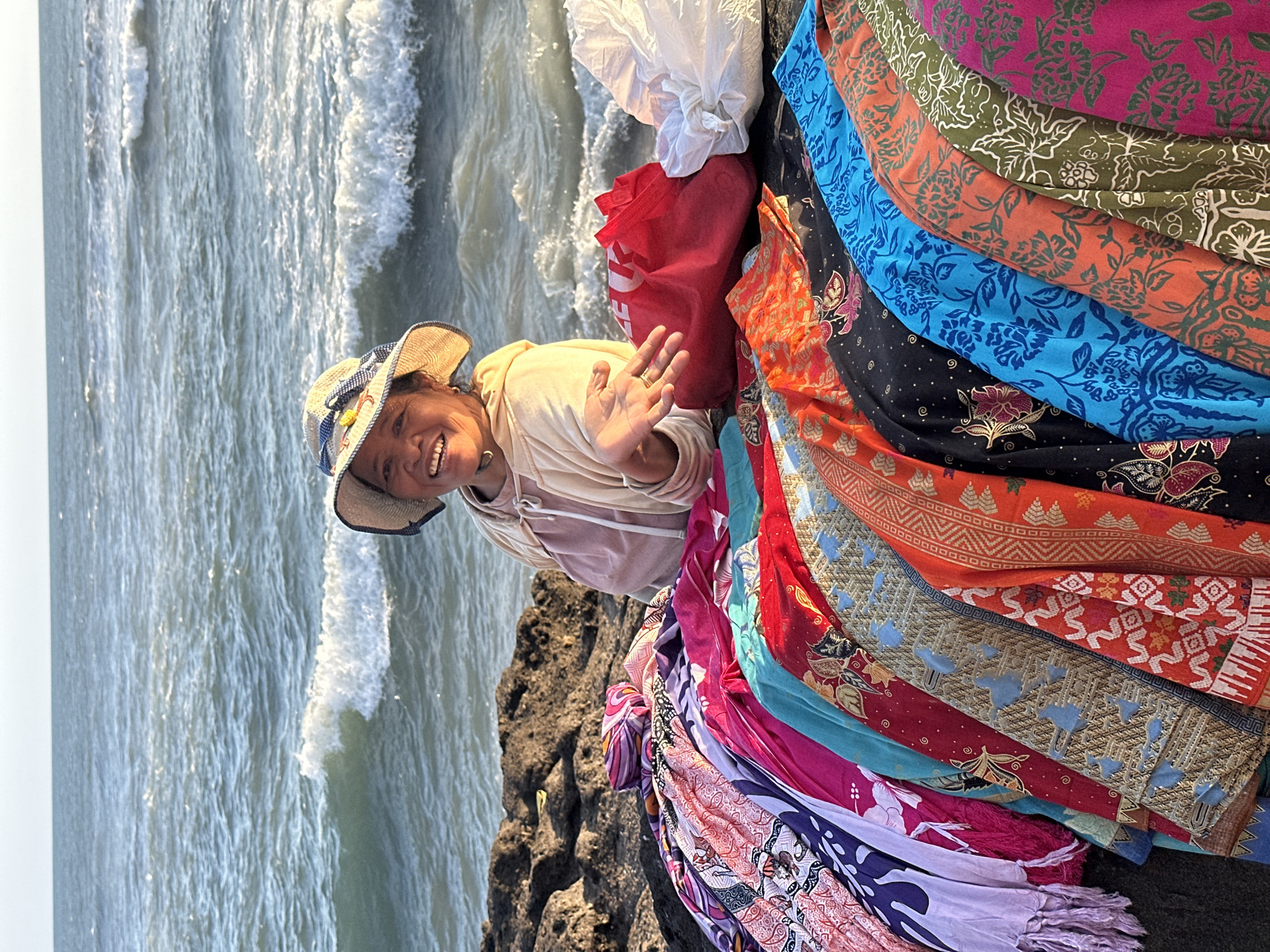 A woman in a straw hat smiles at the camera, gesturing with her open right hand. She is seated behind a display of colorful, patterned fabrics spread across dark, rocky terrain. Behind her, the ocean waves crash against the rocks. The scene appears to be a beachside market in the city of North Kuta, Indonesia, likely in the afternoon due to the lighting. The woman wears a light-colored hooded sweatshirt over a pink garment and a straw hat adorned with a blue and white ribbon and a yellow embellishment. A red tote bag and a white plastic bag are positioned behind the fabrics. The fabrics themselves are richly decorated with various batik-style patterns in vibrant colors including purple, pink, blue, red, orange, and green. The ocean stretches to the horizon under a bright sky.