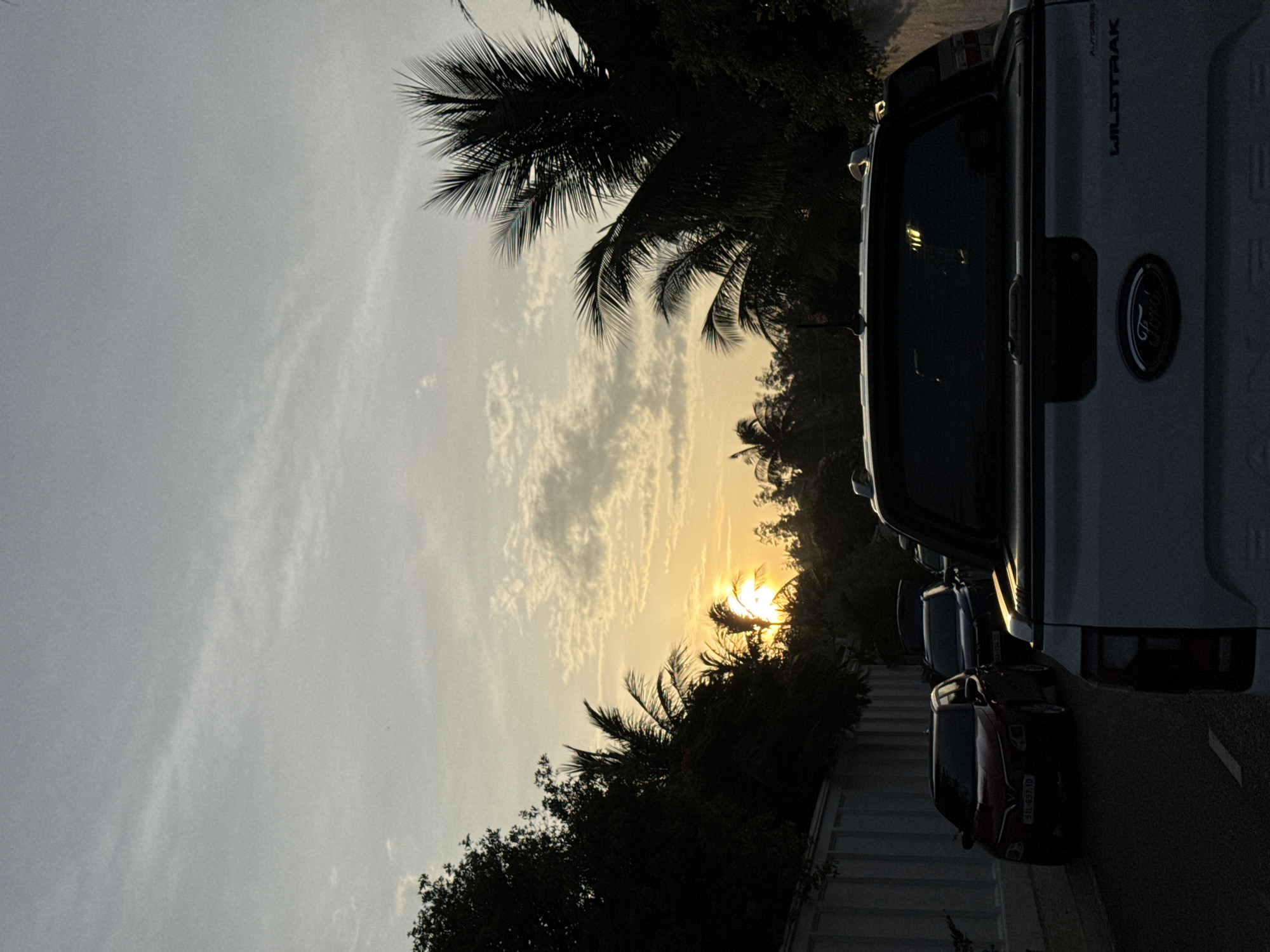 The image depicts a street scene in Phan Thiet, Vietnam, during sunset. In the foreground, the rear of a white Ford Ranger pickup truck is visible, with its "Wildtrak" badging clear on the tailgate. Further down the road, a red sedan is parked, its license plate reading "51L-637.10". Several other vehicles are lined up behind it, indicating traffic. The background is dominated by lush, dark green trees and palm trees silhouetted against a sky illuminated by the setting sun, which creates a soft, warm glow and a scattering of clouds. The overall impression is one of a serene evening commute or a pause in traffic amidst a tropical setting.
