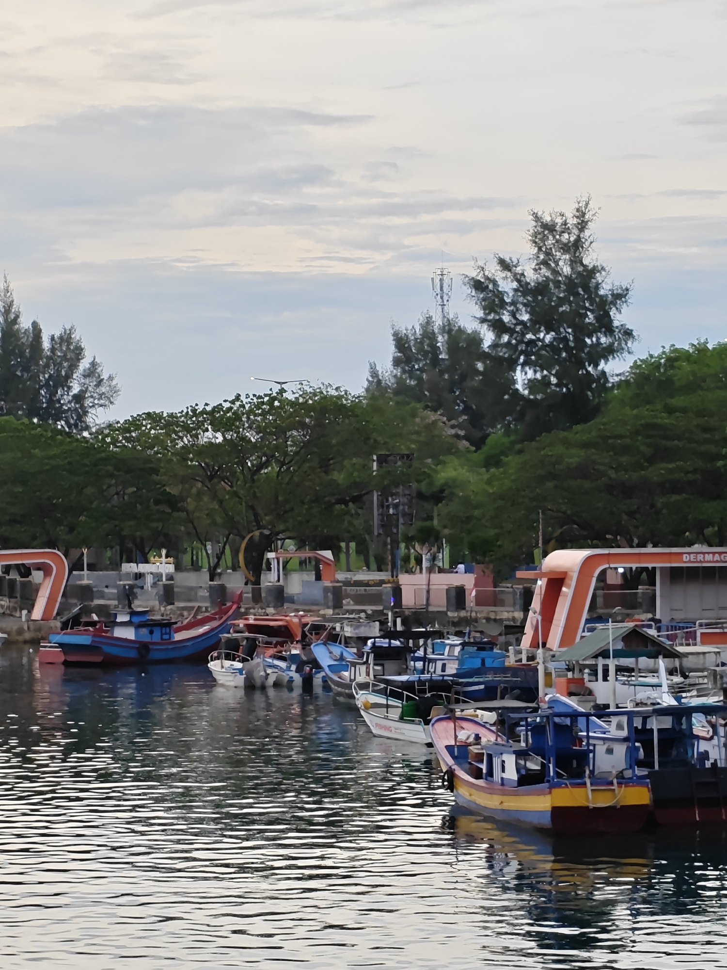 The image shows a harbor in Banda Aceh, Indonesia, filled with small fishing boats. The boats are painted in various colors, including blue, red, yellow, and white. The water in the harbor is calm and reflects the boats and the sky. The shoreline is lined with trees and buildings. A cell tower is visible in the background.