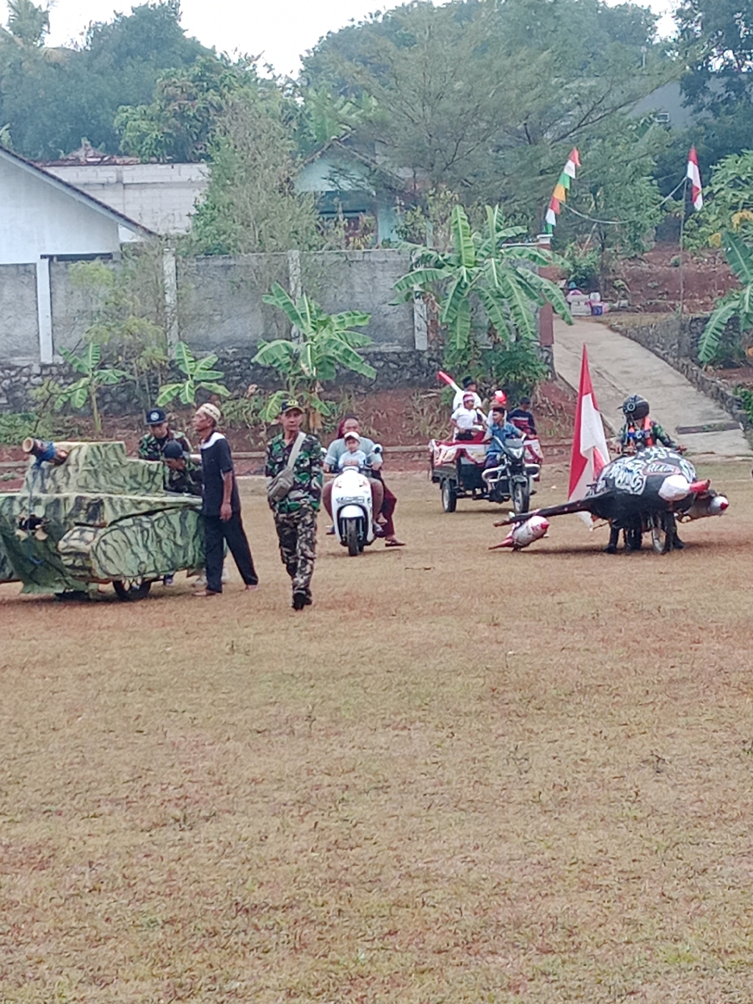 This image captures an outdoor scene in Bojongmengger, Indonesia, likely a public event or festival. In the foreground, a tan-colored grass field stretches out, with a slight incline towards the background. On the left side of the field sits a camouflaged, tank-shaped float, appearing to be made of wood or similar materials, with a visible barrel.

Several individuals are present. A man in camouflage uniform is walking from right to left in the mid-foreground, looking forward. To his left, near the tank float, two individuals in similar camouflage are visible. Another man in a black shirt and a white cap is standing near the tank.

In the middle ground, a white scooter with a rider and passenger moves from left to right. Behind the scooter, a cart is being pulled, with at least three people riding in it. Further to the right, a larger, dark-colored float resembling a creature with horns and a long snout is on display, decorated with a red and white flag. Another small, red object, possibly a projectile, lies on the ground in front of this float.

The background reveals a residential area with buildings and trees. A light-colored house with a dark roof is visible on the far left. A paved walkway leads towards the background, bordered by plants and a stone wall. Two Indonesian flags, one with red and white stripes and another smaller one with multiple colorful stripes, are flown from poles in the background. The sky is overcast, suggesting a cloudy day. The overall atmosphere appears festive and communal.