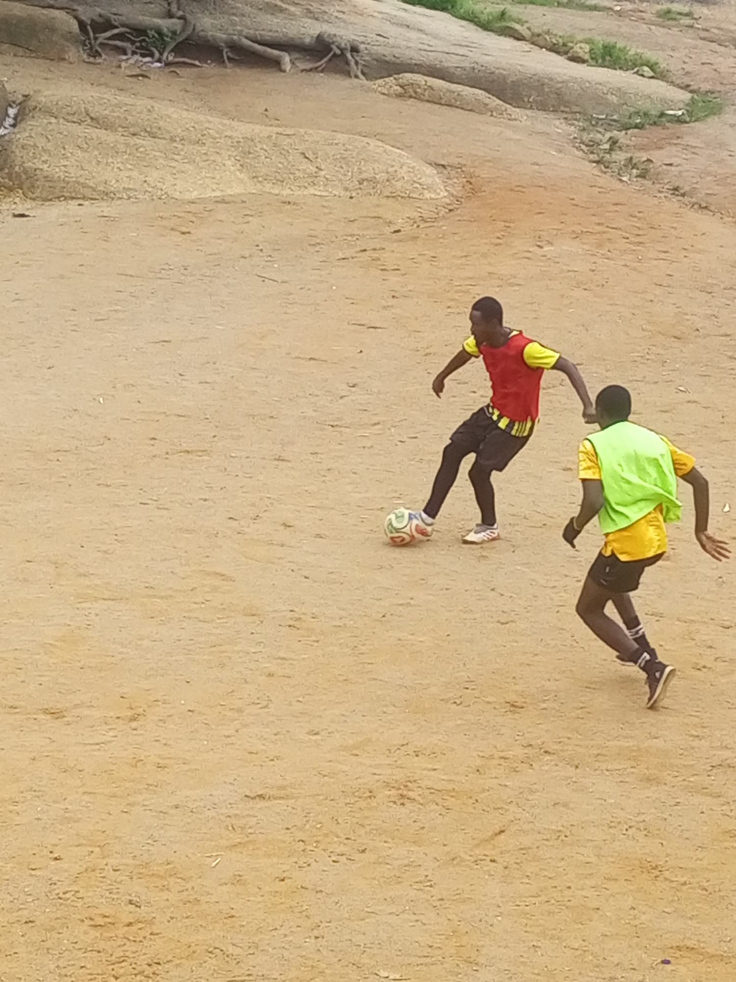 Two young men are playing soccer on a dirt field in Jos, Nigeria. One man, wearing a red and yellow jersey and black shorts, is dribbling the ball. The other man, wearing a yellow shirt and green vest, is defending. The field is surrounded by rocky terrain with sparse vegetation. The overall scene is one of casual athletic activity in an outdoor, natural environment.