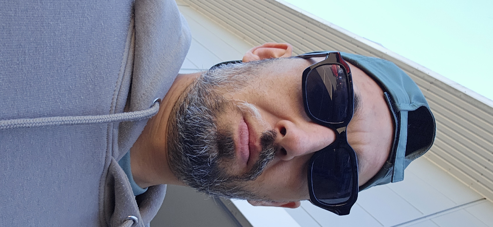 A close-up, low-angle portrait features a man with a salt-and-pepper beard and mustache, looking slightly upwards. He is wearing dark, rectangular sunglasses that cover his eyes. On his head, he wears a dark green baseball cap, positioned with the brim facing backward. He is dressed in a light grey hooded sweatshirt, the fabric of which shows some pilling, and a grey drawstring is visible across the front. A hint of a light blue garment can be seen under the collar of his hoodie.
The background consists of light-colored, horizontally paneled siding of a building on the left and a clear, bright blue sky in the upper right. The bright lighting suggests it is daytime with good weather. There is no visible text in the image. The man's expression is obscured by the sunglasses, and no specific activity is taking place other than posing for the photograph. The setting appears to be outdoors next to a building, possibly in Norwich, United Kingdom as per the provided context.