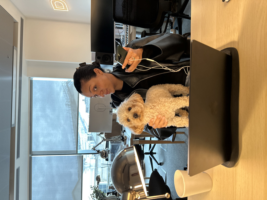 An adult female, with dark hair pulled back, wearing a black leather jacket over a black top, is seated at a light-colored wooden desk in an office environment. She holds a small, light-brown, curly-haired poodle-type dog with her right arm, while simultaneously holding a dark-colored smartphone with white wired headphones plugged in, in her left hand. The dog stands upright, facing the camera. The woman looks directly at the camera with a slight smirk and raised eyebrows.
On the desk in the foreground, a black laptop rests on a stand, open and facing away from the viewer. To the left of the laptop is a white mug and an illuminated chrome desk lamp. The background reveals an open-plan office space featuring additional desks, ergonomic chairs, computer monitors (one displaying a white "O" or ring-shaped graphic), studio speakers, and more chrome desk lamps. Large windows, partially covered by blue roller blinds, form the rear wall. Through the blinds, various urban structures are visible, including what appears to be the upper part of a classical building with distinct railings, resembling the Royal Courts of Justice. This architectural detail places the location in the vicinity of Fleet Street or the Strand, in the City of London, United Kingdom. A subtle neon sign displaying a stylized letter "D" is visible on the far right background wall.