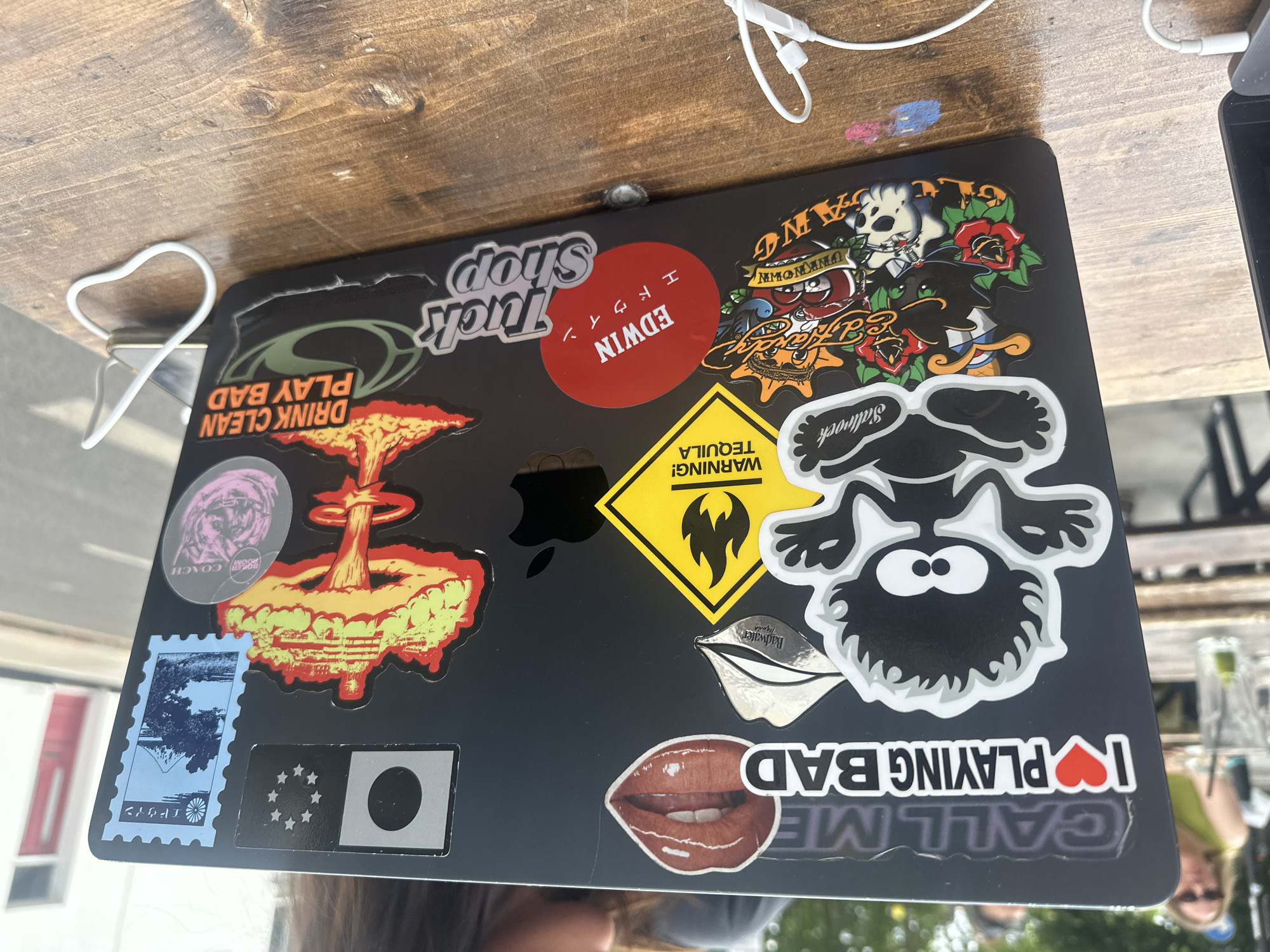 The image shows a black laptop covered in various stickers. The laptop is on a wooden table, with a pair of white earphones nearby. Stickers include brands like "Edwin," "Saltrock," and "Badwater," alongside images like a skull, a mushroom cloud, lips, and a warning sign for tequila. The overall aesthetic suggests a casual, personalized style. The location in London, United Kingdom is not evident from the image.