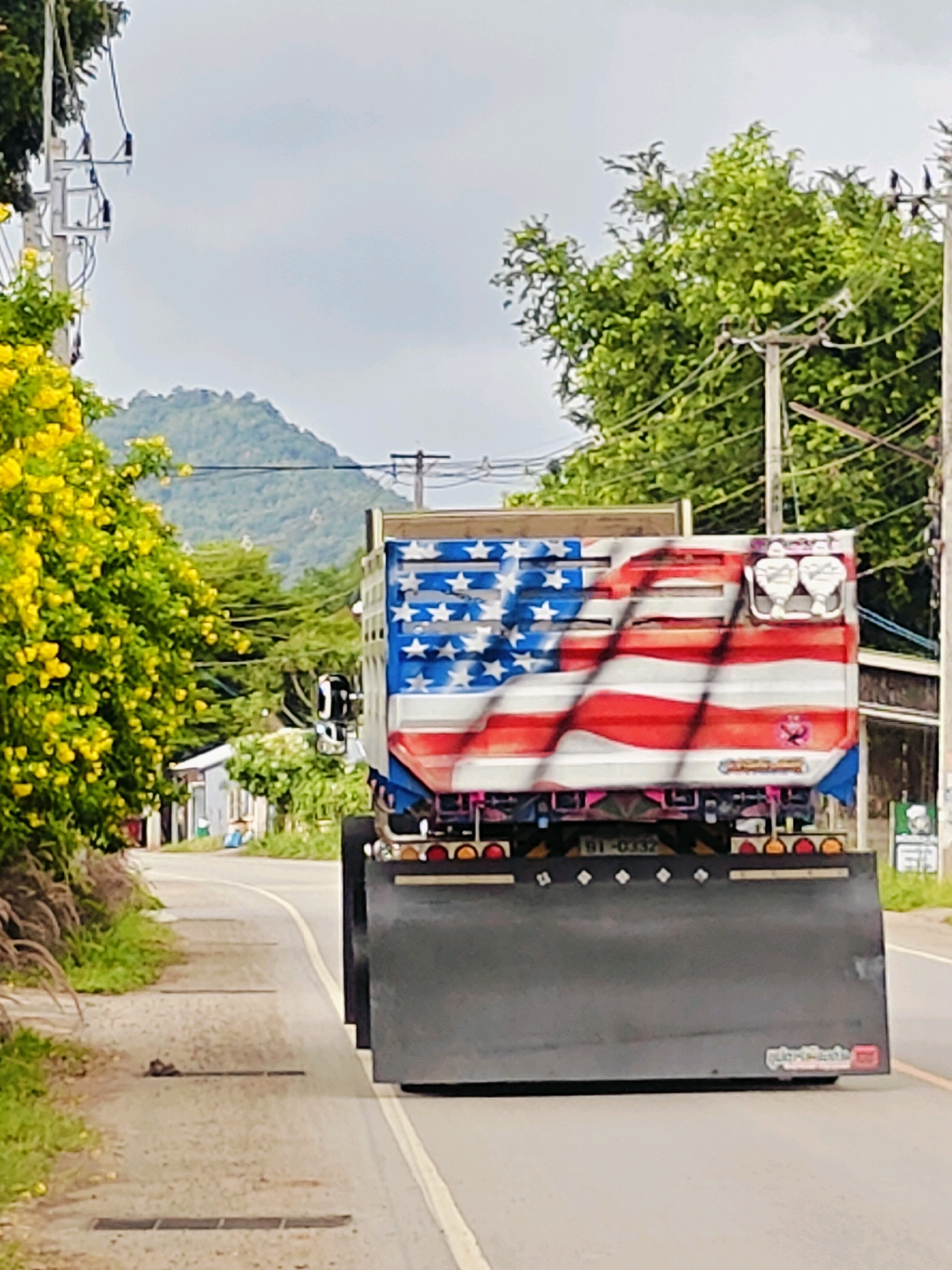The image shows the rear of a dump truck with an American flag design painted on its side, driving on a road in Mu Si, Thailand. The truck has a large, dark gray mud flap. The surrounding environment includes lush green trees, a mountain in the background, a cloudy sky, and utility poles with wires. Yellow flowers are visible on the left side of the road. A license plate on the truck reads "BI-0332".