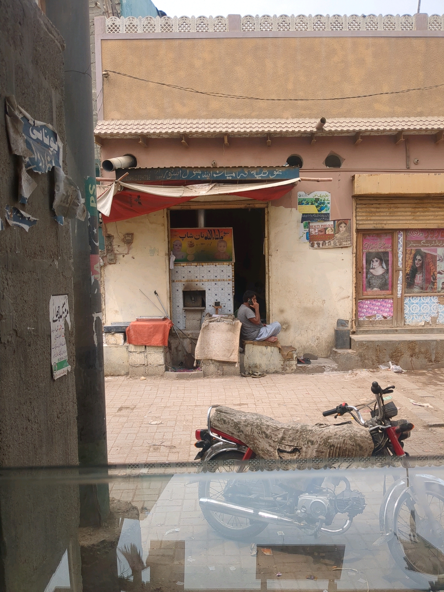 The image captures a street scene in Karachi Division, Pakistan, likely on a sunny day. In the foreground, a motorcycle is partially visible, covered with a camouflage-patterned fabric. Its reflection is seen on a wet surface, possibly a puddle or a wet pavement, which also reflects the surrounding environment.
The main subject of the image is a small shop or business establishment. A man is seated on a raised platform outside the entrance of the shop, talking on a mobile phone. He is wearing a striped shirt and light-colored pants. The shop's facade features a red awning over the entrance and posters displayed on the walls. One poster shows an image of babies, while another advertises "BEAUTY PARLOUR & TRAINING CENTRE" with images of women. The shop's shutters are visible on the right side of the image.
The environment appears to be an urban street with tiled pavement. There are also some discarded items on the ground, suggesting a busy, everyday setting. The building itself has a textured, light-colored facade with decorative elements along the roofline. A pipe protrudes from the building's wall above the awning. Torn posters are affixed to a rough, grey wall on the left side of the image.