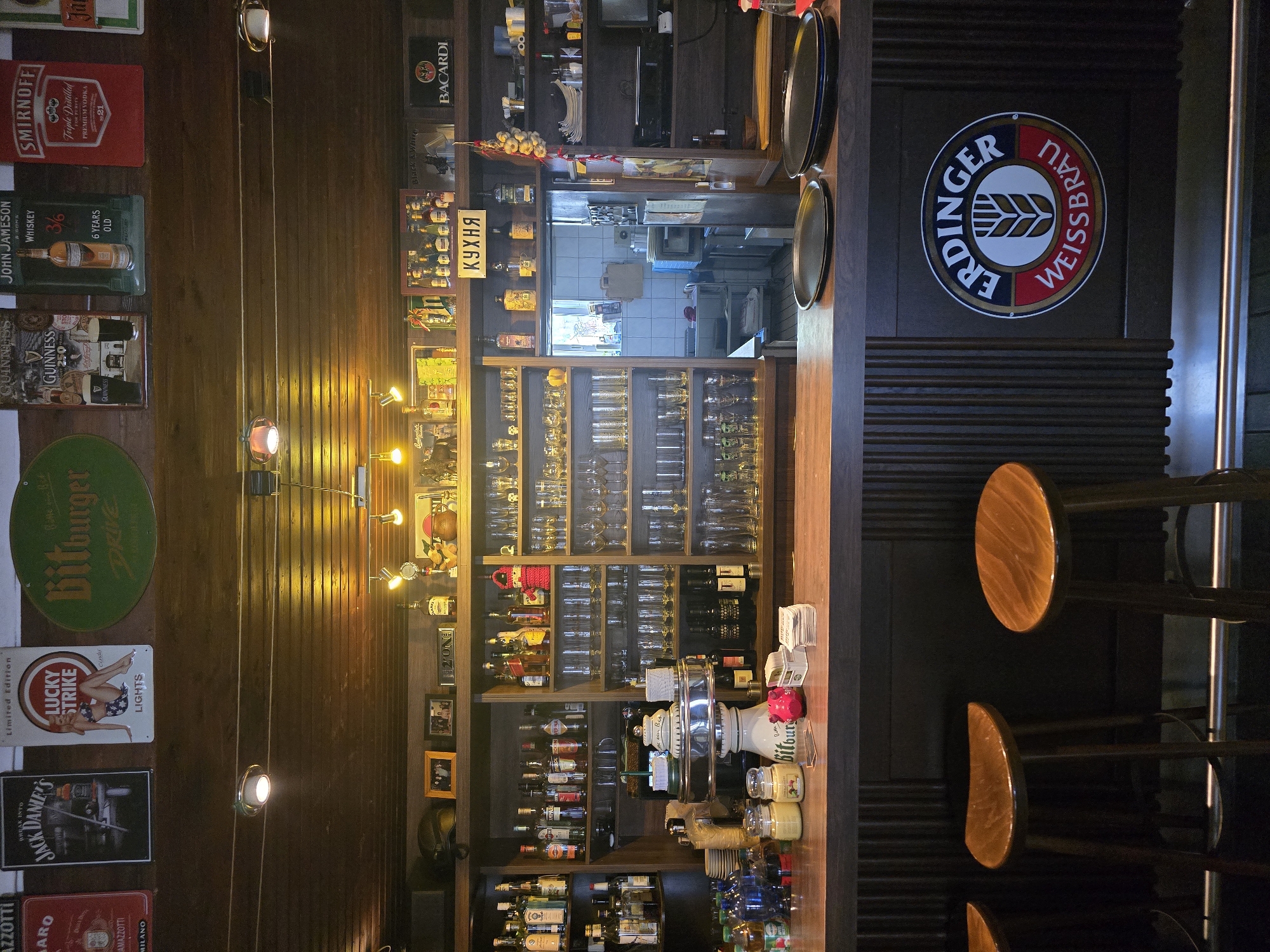 The image shows a well-stocked bar interior, likely in Saarbrücken, Germany, based on the presence of the Erdinger Weißbräu logo prominently displayed on the bar front. The bar area is characterized by dark wood paneling on the walls and ceiling, creating a cozy, traditional atmosphere.
The top shelf displays a collection of vintage-style signs and advertisements, including Jack Daniel's, Lucky Strike, Bitburger Drive, Guinness, John Jameson, and Smirnoff. Below this, a second shelf holds various bottles of alcohol and some decorative items.
The main bar counter is made of dark wood, with various bottles, jars, and barware arranged on it. Notably, there is a beer tap system with a chrome finish. Behind the counter, a large shelving unit extends upwards, filled with an extensive collection of glasses of various types and sizes, as well as more bottles of spirits. To the right of the shelves, a small kitchen or service area is visible through an open doorway.
In the foreground, three wooden bar stools are positioned in front of the counter. On the bar itself, two dark circular platters are placed. The overall scene suggests a functional and inviting establishment, likely a pub or a bar, that caters to patrons looking for a wide selection of beverages in a classic setting. There are no people visible in the image.