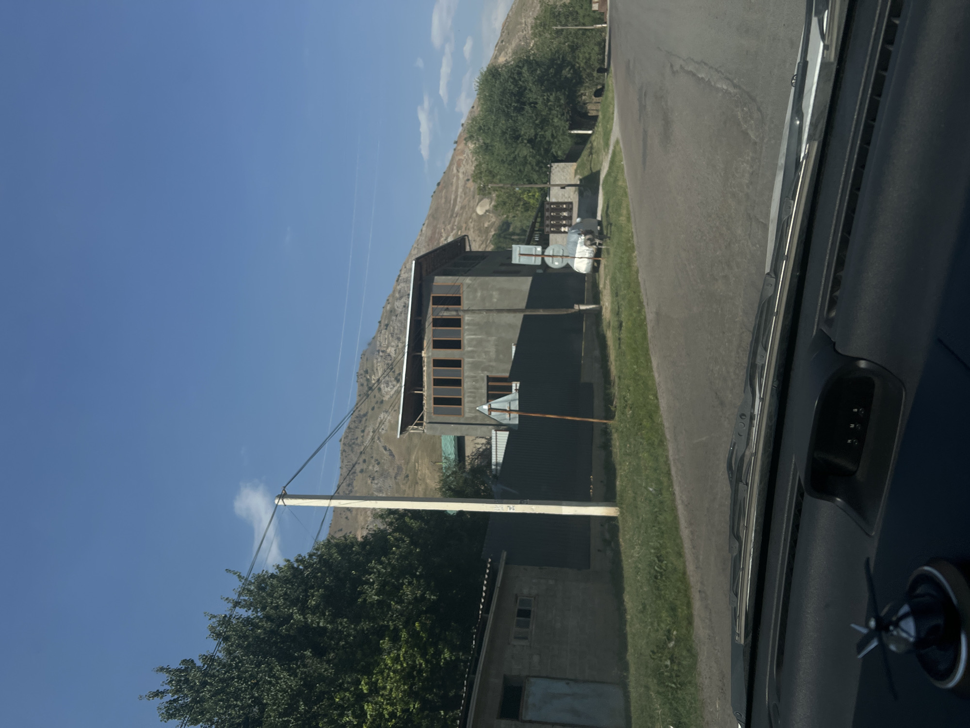 A residential street in Yakkatut, Uzbekistan, is depicted on a sunny day. The scene is viewed from inside a vehicle, looking forward through the windshield. On the left side of the road, there is a single-story brick building with a metal door and windows. Adjacent to it is a dark green metal fence. A tall utility pole stands in front of the fence, with electrical wires running across the frame. Beyond the fence, a large, multi-story building with a prominent upper floor featuring large, multi-pane windows is visible. The building appears to be under construction or in a state of renovation, with exposed rafters on the roof. In the background, a steep, arid hillside dominates the landscape. To the right of the road, there are trees and another smaller building, as well as a white car covered with a tarp. The road itself is paved and appears to be in moderate condition. The sky is clear blue with a few wispy clouds. The dashboard of the vehicle is visible in the foreground, with a decorative item on the left and a small electronic display on the right.