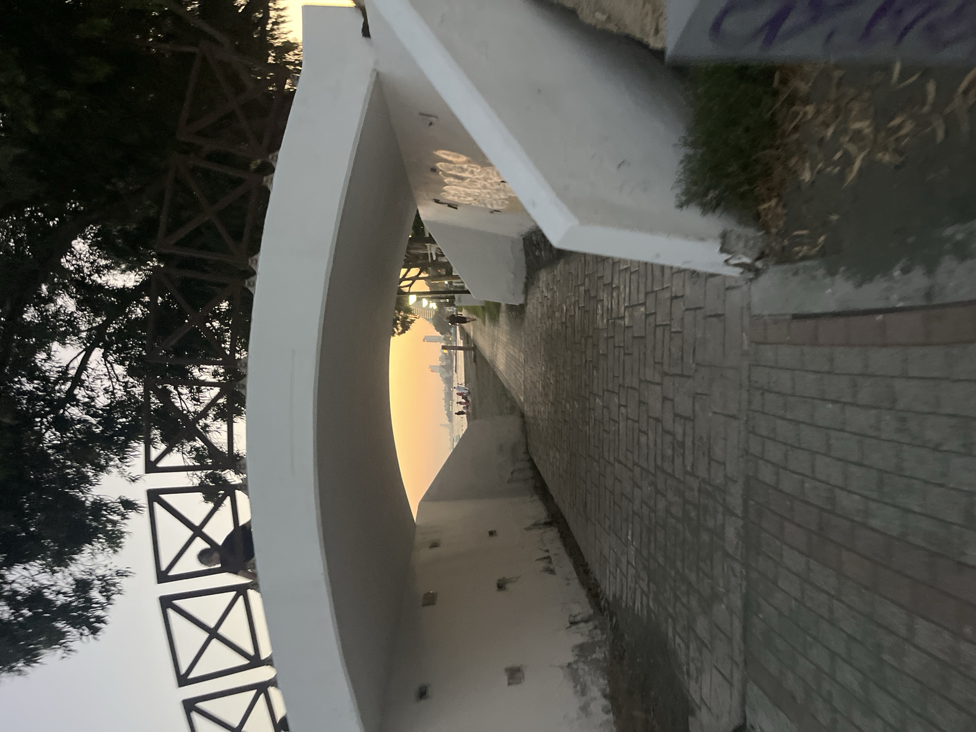 A pedestrian walkway, paved with rectangular stones, leads under a white arched bridge. The walkway is bordered by a white wall on the left and a sloping white structure on the right, which has graffiti on it. Beyond the bridge, the walkway opens onto a beach with several people visible. The sea is in the background, with buildings and trees lining the shore. The sky is a warm orange, suggesting it is either sunrise or sunset. A person is sitting on the railing of the bridge, looking out towards the beach. Trees with dark foliage are visible above the bridge. The overall scene depicts a coastal urban environment with a recreational atmosphere.