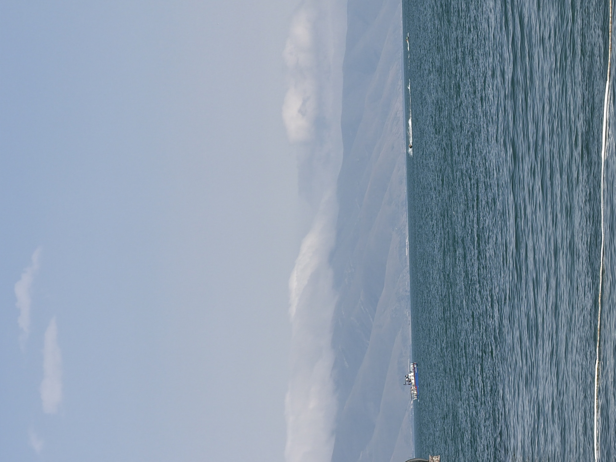 A wide shot captures a tranquil, vast blue body of water under a hazy sky with sparse white clouds. In the distance, a chain of mountainous terrain stretches across the horizon, partially obscured by a low-lying bank of white clouds that follows the contours of the peaks. A small blue and white boat with several people on board is visible on the water to the left of the frame, appearing to be a passenger ferry. To the right, two jet skiers are actively moving across the water, leaving trails of white spray behind them. The foreground of the image is dominated by the rippling surface of the water, with a white rope or line visible near the bottom edge, partially submerged. The overall impression is one of a leisurely day on the water, with recreational activities taking place against a scenic, natural backdrop. The location is the Sevan lake, near the city of Tsamakaberd in Armenia.