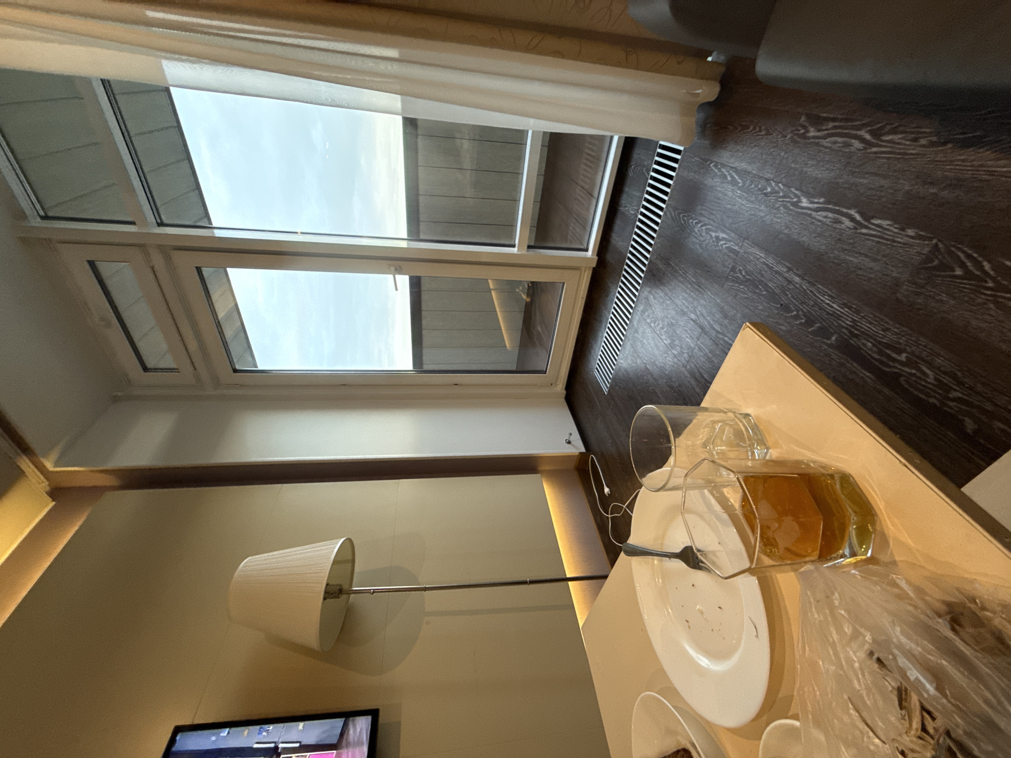 The image shows the interior of a room in Dom Otdykha Zolotoy Plyazh, Russia. A double-paned window is visible, offering a view of what appears to be another building. A floor lamp with a white shade stands in the corner of the room, its metal rod extending towards a side table. On the table are a partially empty glass, a used plate with a fork, and discarded food packaging. The floor is made of dark brown wood planks with a linear grate near the window. A television screen is seen in the lower left corner of the image. The walls are light beige with a decorative gold trim along the top.