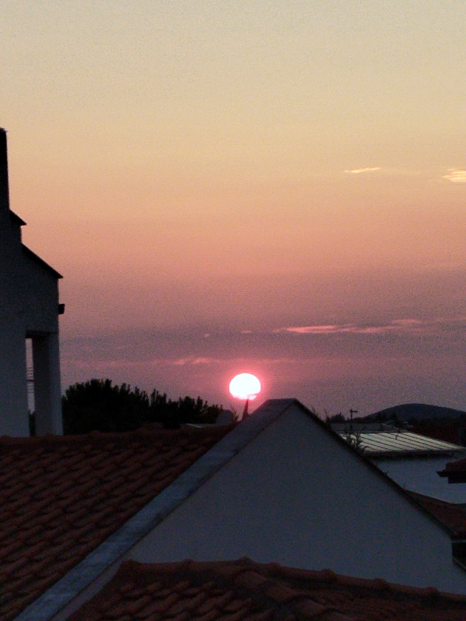 An outdoor, low-angle, medium shot captures a sunset over a residential area in Sithonia Municipal Unit, Greece. The sky is a gradient of pale yellow at the top, transitioning to pink and then to a deep purple. The sun, a bright pink circle, is partially obscured by thin clouds and appears low on the horizon, casting a reddish glow. In the foreground, terracotta tiled rooftops dominate the bottom half of the frame, with the dark peak of a modern building with white walls visible on the left. Behind the rooftops, a silhouette of dense green trees is seen against the sky, with a dark hill or mountain range in the far background. The scene appears to be devoid of people, and the overall atmosphere is tranquil and serene.