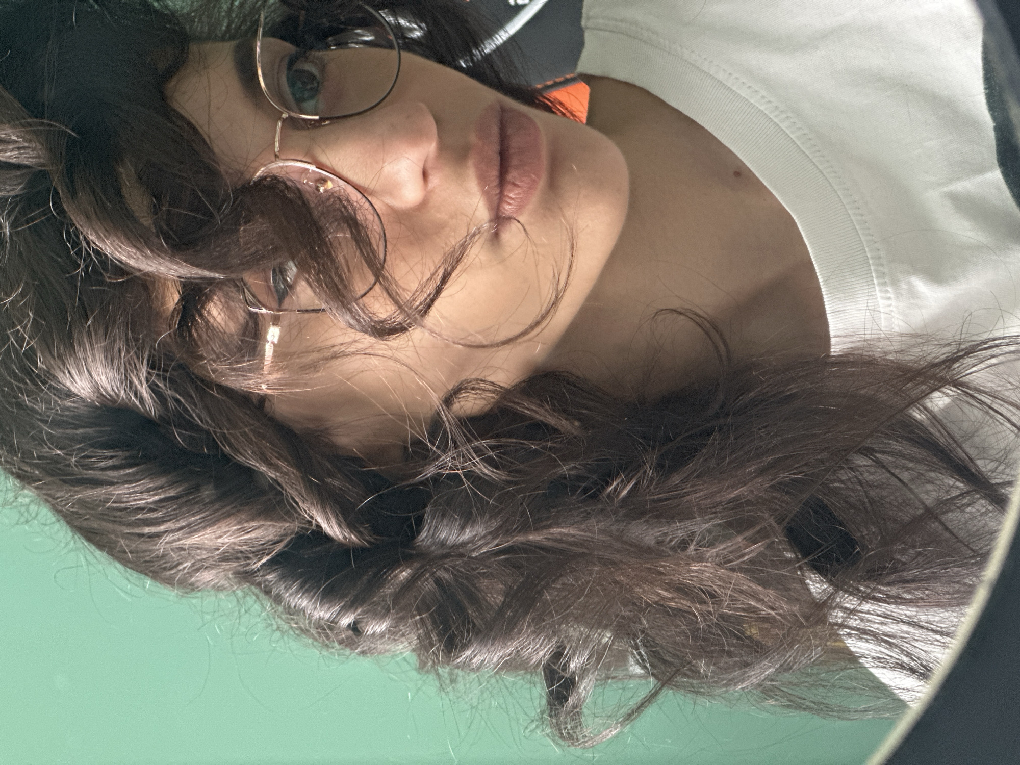 The image shows a close-up, head-and-shoulders portrait of a young woman with dark, curly hair and glasses. She is wearing a white t-shirt. The background is a solid, muted green color. The lighting is bright, creating highlights on her hair and skin. The woman is looking slightly to the side, and her lips are parted. The image appears to be a selfie, as evidenced by the partial reflection of what seems to be a phone or camera in the lower left corner of the frame. The scene is indoors, with no discernible features of a city, let alone Sofia, Bulgaria.