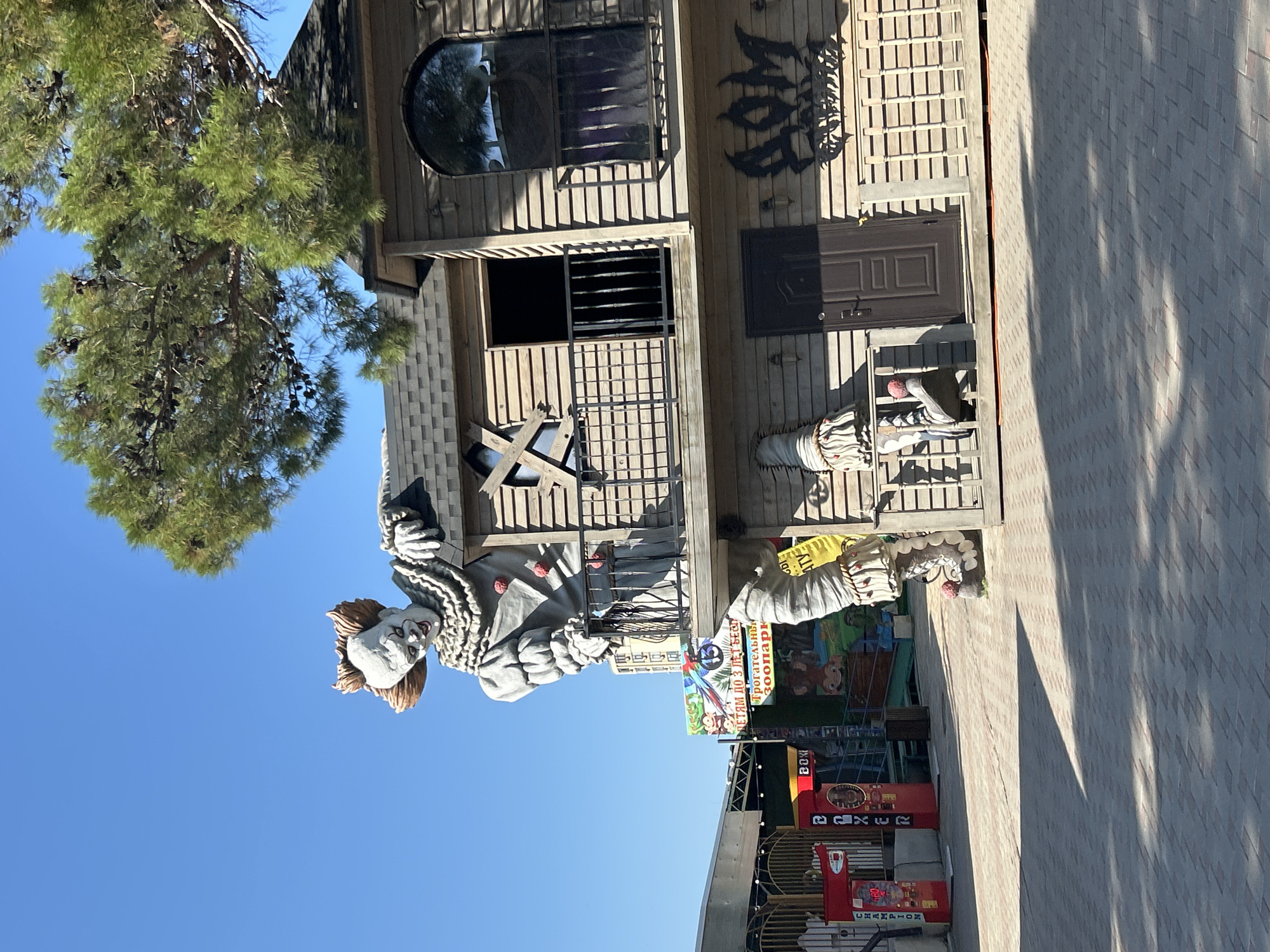 The image depicts a peculiar building in Gelendzhik, Russia, designed to resemble a haunted house. Dominating the scene is a large, grotesque statue of a clown, reminiscent of Pennywise from Stephen King's "It," perched atop the roof. The clown figure is positioned as if peering over the edge of the roof, with its arms resting on the building's structure.
The building itself is constructed from wooden planks and features a two-story design. It has balconies with black metal railings and arched windows. One window is boarded up with wooden planks forming an 'X'. The lower part of the building shows what appear to be the legs and feet of the clown figure emerging from the structure, further enhancing the unsettling aesthetic. A dark brown door is visible on the lower level, and above it, graffiti in a stylized font reads "ДОМ," which translates to "house" in Russian.
In the foreground, a paved area is visible, with shadows cast by an unseen object, likely a tree, as indicated by the pine branches extending into the upper right corner of the frame. To the left of the main building, there are some arcade-style game machines, including a "BOXER" machine. A banner with Cyrillic text and cartoon animals, including a parrot and monkeys, is also visible, suggesting the location is an amusement park or entertainment venue. The sky is a clear, bright blue.