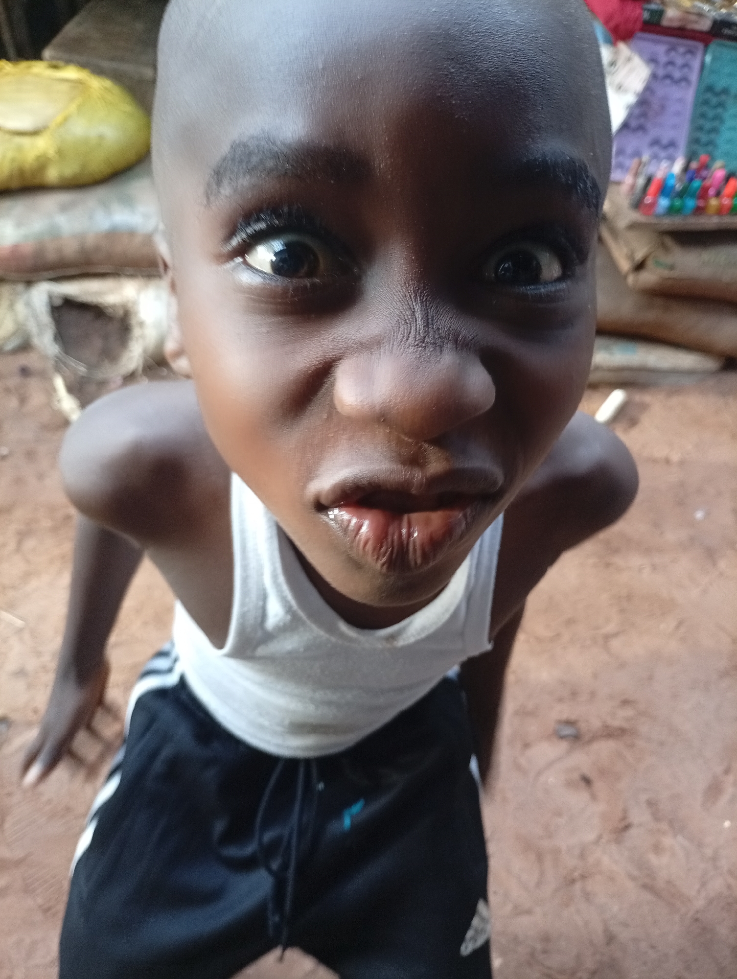 The image shows a close-up portrait of a young Black boy in Nkpor, Nigeria. He is looking directly at the camera with a playful expression, his eyes wide and his lips slightly parted. He is wearing a white sleeveless shirt and black shorts with white stripes. The background is blurred, but appears to be an outdoor setting with reddish-brown earth, possibly a dirt path or courtyard. There are some bags and colorful items visible in the background, suggesting a market or a home environment. The lighting indicates it is daytime. The focus is on the boy's face and upper body, with his skin appearing smooth and his hair shaved. The overall impression is one of innocence and curiosity.