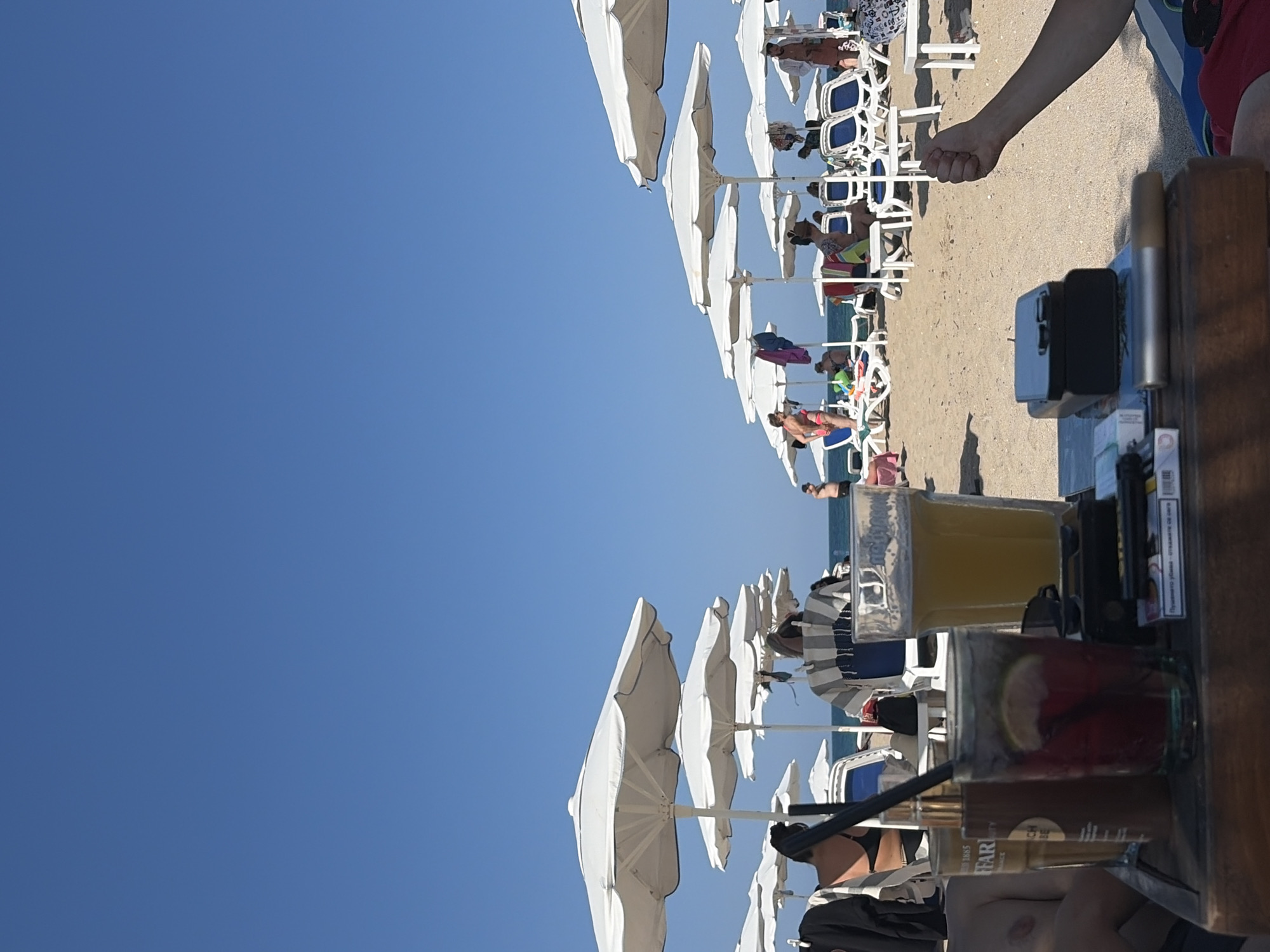 The image depicts a sunny day on a beach in Lozenets, Bulgaria. In the foreground, a wooden table holds two drinks, one a red cocktail with a lime slice, and the other a glass of light-colored liquid, possibly beer. A pack of cigarettes and a black electronic device are also on the table. A person's arm is visible in the lower right corner, with their fist clenched.
Beyond the table, the beach is lined with white umbrellas and beach chairs, many of which are occupied by people. Some individuals are seated, while others are standing or walking. The sea is visible in the background under a clear blue sky. The overall scene conveys a relaxed atmosphere of a beach resort.
