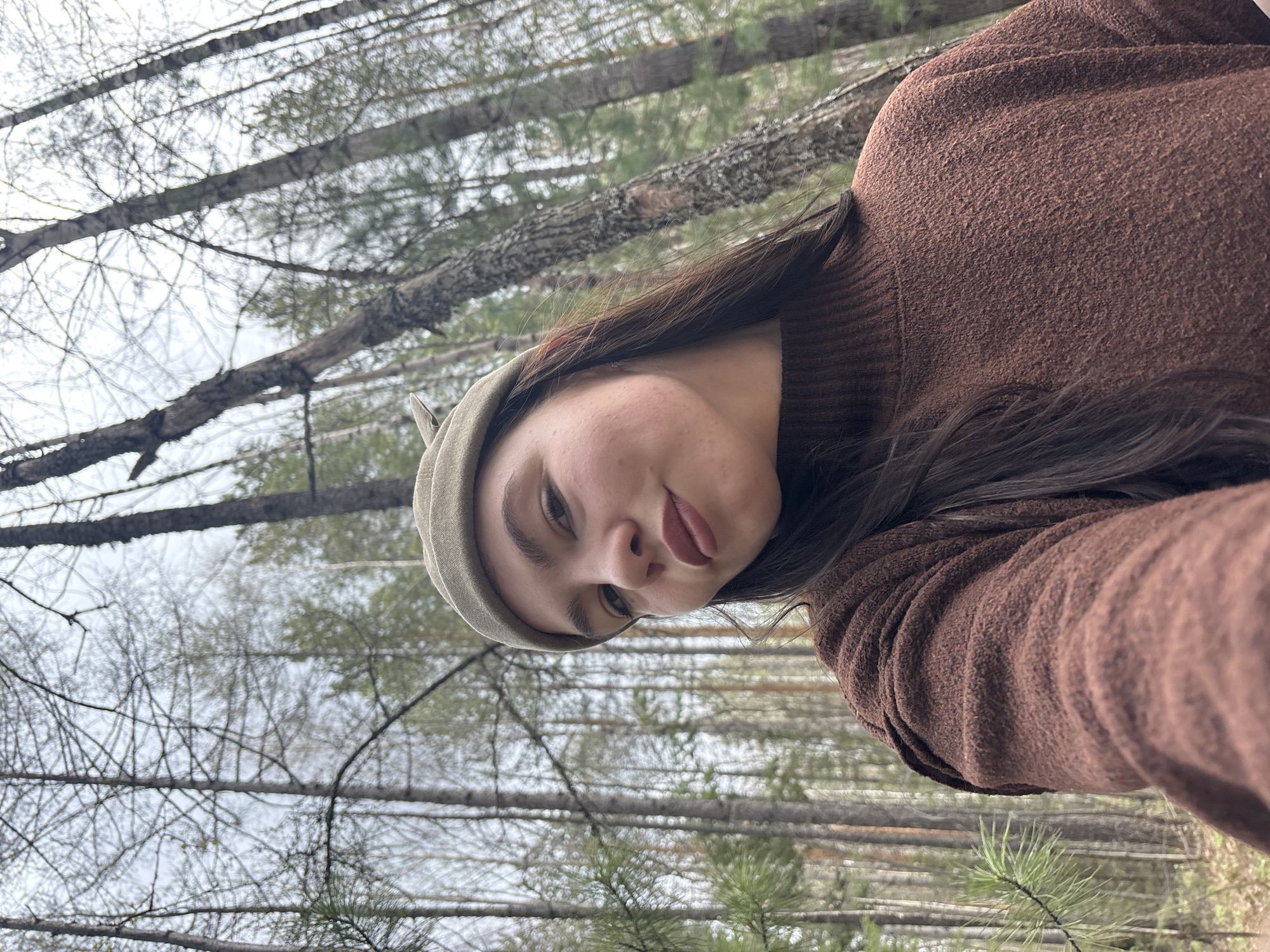 A person is depicted in a selfie format within a wooded area. The subject is wearing a brown, textured sweater and a light green cap. Their hair is dark and falls to the side. The background consists of a forest setting, with numerous trees exhibiting bare branches against an overcast sky. The location is near Kachkanar, Russia, as the trees appear similar to that of a birch forest. A single pine branch is visible in the lower center of the frame.