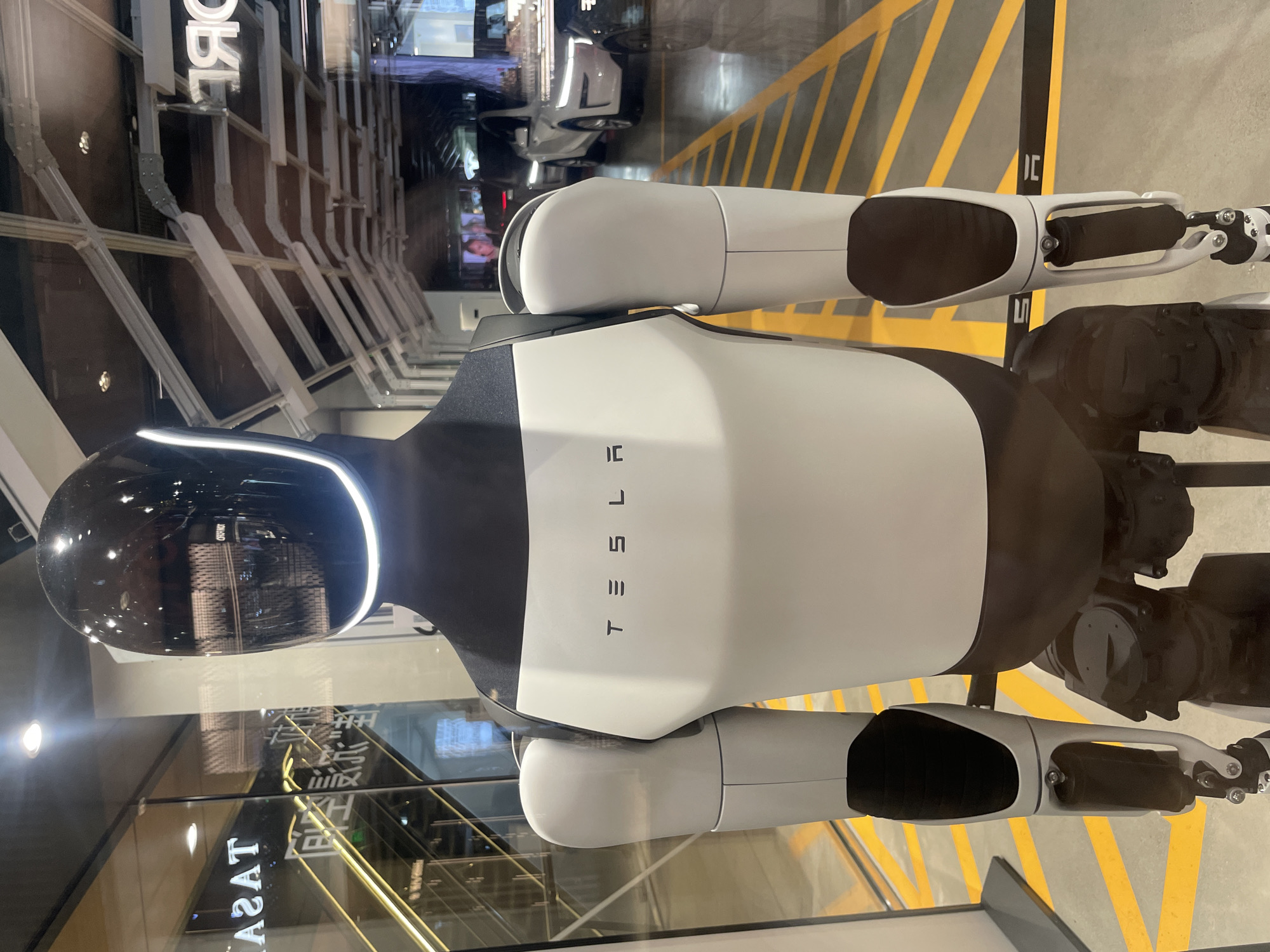 A Tesla Optimus robot is displayed indoors behind glass. The robot has a white torso with "TESLA" printed vertically in black lettering. It has a black head and shoulders. The arms are predominantly white with black grips. A strip of light is visible around the head. The background shows reflections of the surrounding environment, including a store, scaffolding, and yellow stripes on a surface, possibly indicating a road or parking area. The location is inferred to be in Jinjiang District, China, based on possible Chinese characters in the reflection.
