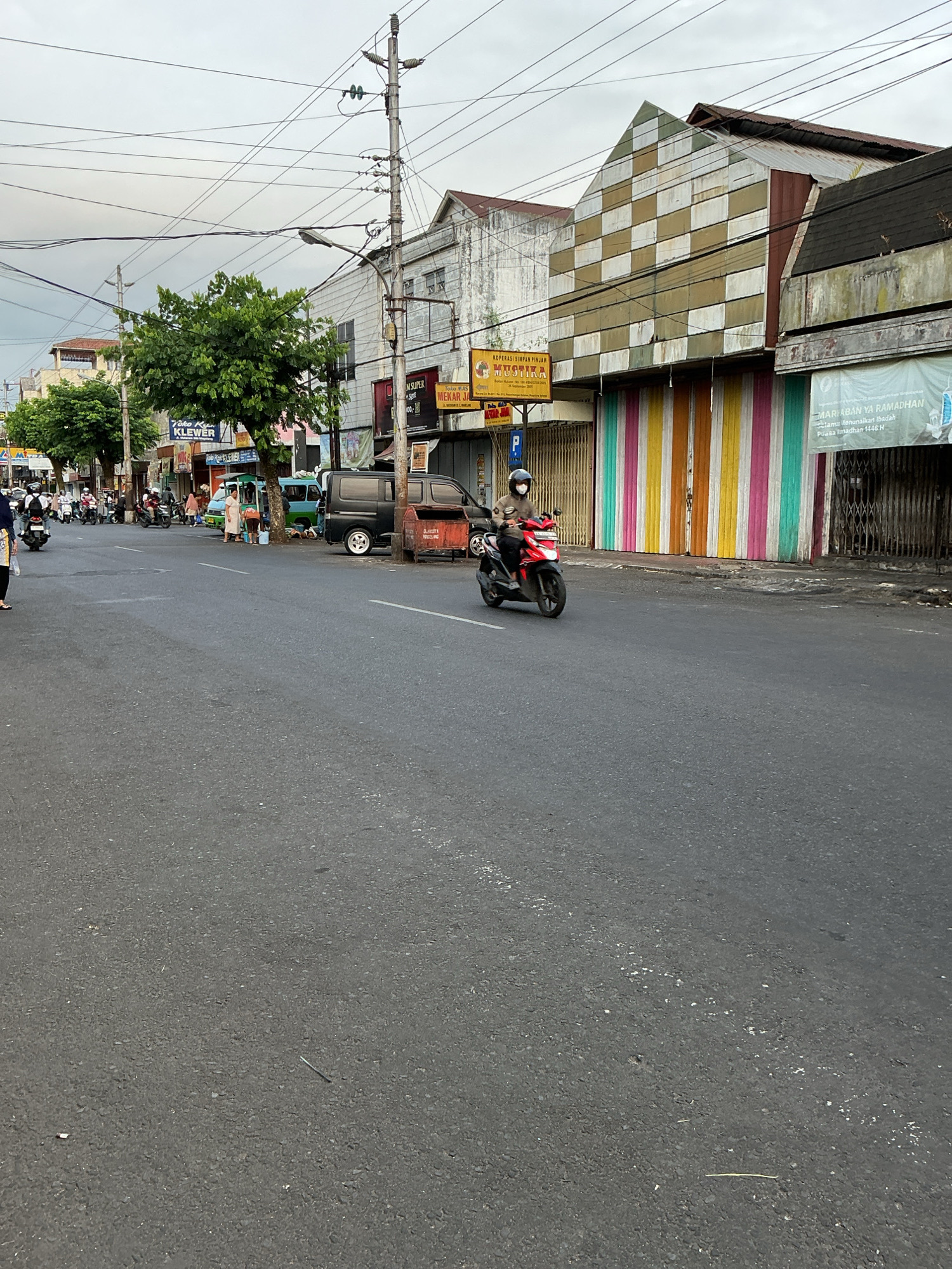 The image shows a street scene in Magelang, Indonesia. A person is riding a red motorcycle on the right side of the road, wearing a helmet and a brown jacket. The road is asphalt and appears to be a two-lane street. On the left side of the street, there are several buildings with shopfronts, including one with a sign that reads "Toko Kiu KLEWER." There are also several trees and utility poles with many wires running between them. In the background, there are more buildings and a cloudy sky. The buildings on the right side of the street have colorful metal shutters. A banner with Indonesian text that mentions "RAMADHAN" is visible on one of the buildings. There are other vehicles and pedestrians on the street, indicating a normal day of activity.
