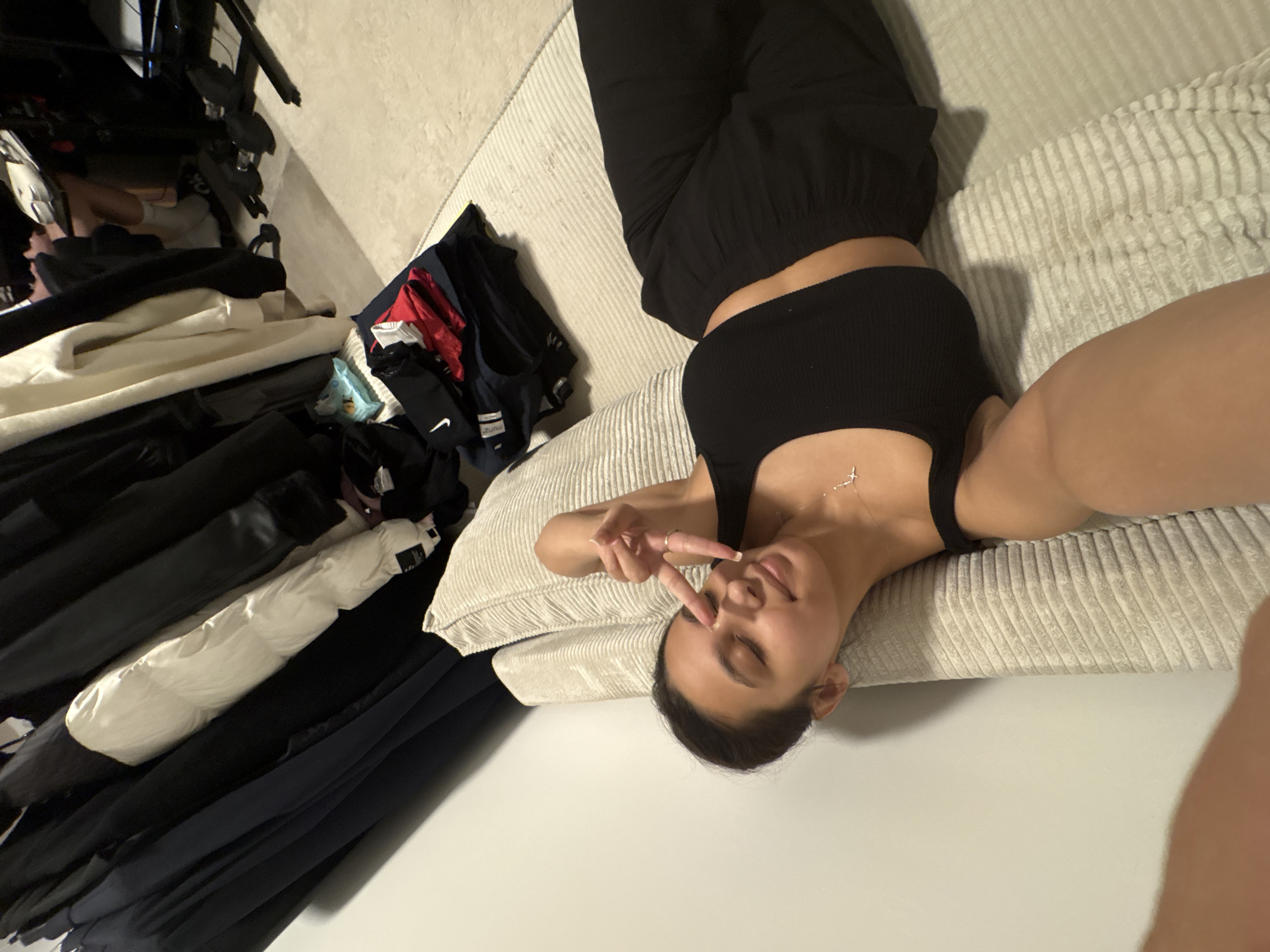 A young woman is depicted lying on her back on a light-colored, ribbed sofa or bed, posing for a photo. Her head rests on a matching pillow, and her eyes are closed, conveying a serene or playful expression. She is making a peace sign with the fingers of her right hand, which is positioned near her face. She wears a black ribbed crop top and black pants, and a delicate silver cross pendant necklace is visible around her neck.
The scene is indoors, likely illuminated by artificial light. To the upper left of the woman, a cluttered area is visible, dominated by a large pile of clothes. Garments in black, white, dark blue, and red are stacked or draped, suggesting a casual, lived-in environment. A white Nike swoosh logo is clearly visible on a dark piece of clothing within this pile. Another piece of clothing appears to have the text "ZUNU". Among the jumbled items, a white puffy jacket or garment stands out. The overall impression is a relaxed moment captured in an informal, possibly domestic, setting.