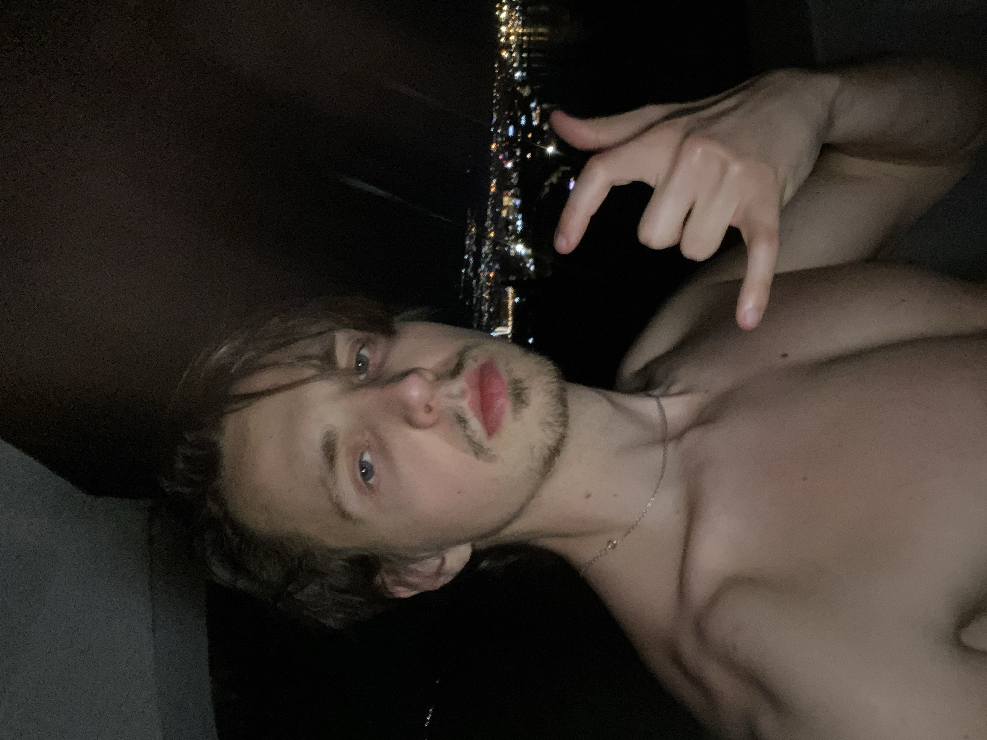 A shirtless man with light-colored hair and blue eyes is posing for a selfie at night in Šušanj, Montenegro. He is making a hand gesture with his right hand. The lights of the city are visible in the background.