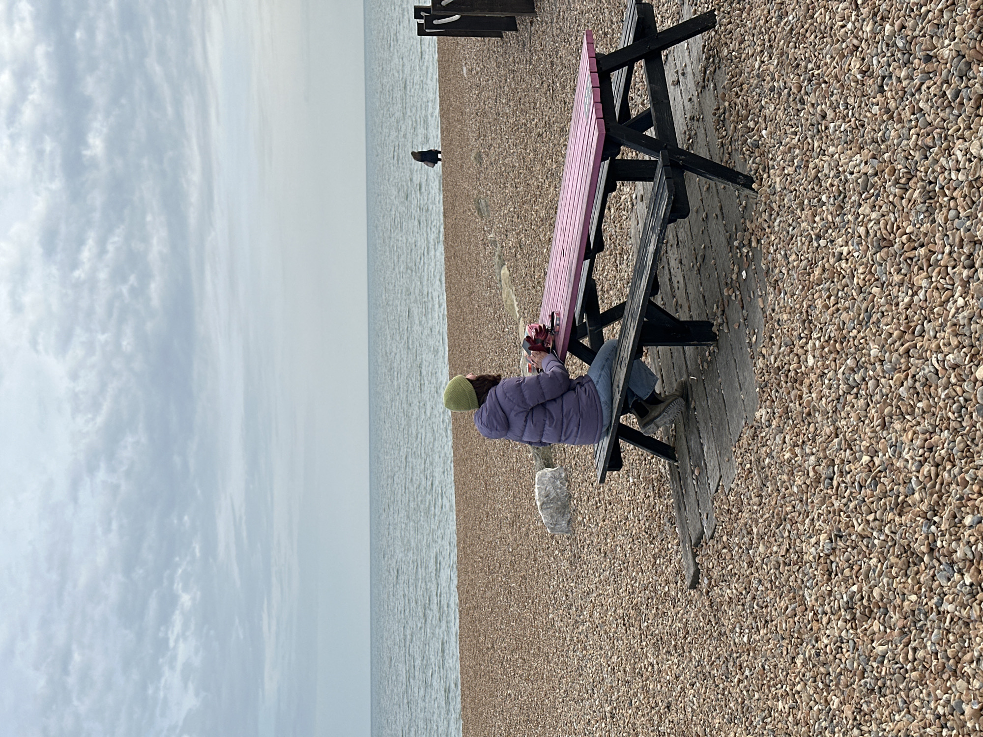 A woman sits on a wooden bench at a pebbled beach in Folkestone and Hythe District, United Kingdom. She wears a lilac jacket and a green beanie. She appears to be interacting with objects on the pink painted table of the bench. In the background, the calm sea meets the horizon under a cloudy sky. A single, distant figure stands near the water's edge.