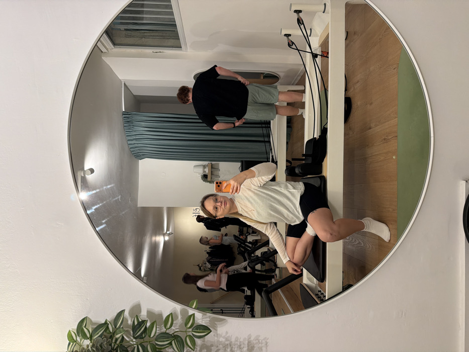 A woman captures a mirror selfie in what appears to be a fitness or pilates studio, located in Tel-Aviv, Israel. The photo, initially rotated 90 degrees counter-clockwise, shows the woman with blonde hair and glasses, wearing a light-colored long-sleeved top, dark shorts, and white socks. She is kneeling on a white pilates reformer, holding an orange-cased smartphone to take the picture, looking directly into the large circular mirror with a thin white frame.
In the reflection of the mirror, behind the woman, a man with reddish-brown hair is visible, dressed in a black t-shirt and light-colored shorts, standing with his hands on his hips. Further in the background of the reflection, several other people can be seen, likely also exercising on various pieces of equipment, possibly spin bikes. The studio features wooden flooring, white walls, and dark teal or blue-green curtains covering a section of the wall. Artificial lighting fixtures are visible on the walls, and a green exercise mat is present on the floor near the reformer. A small shelf with a decorative item is also reflected on the back wall. In the actual room, outside the mirror's frame, the top of a leafy green plant is visible in the foreground on the left. The overall setting is clean and modern. No discernible text is visible in the image.